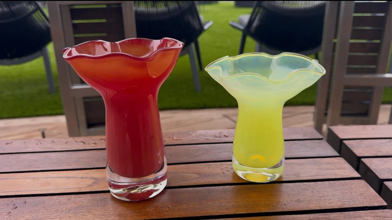 Two colorful glass vases with wavy, flower-like rims on a wooden table outside. One vase is red, the other is yellow, and both are empty.
