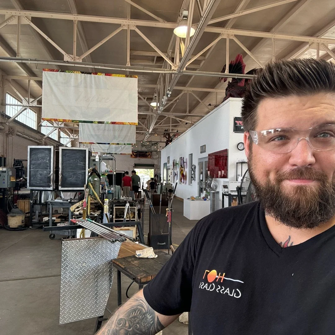 A bearded man with glasses taking a selfie in a large industrial-style workshop or studio with high ceilings, bright lighting, and various tools and equipment in the background.