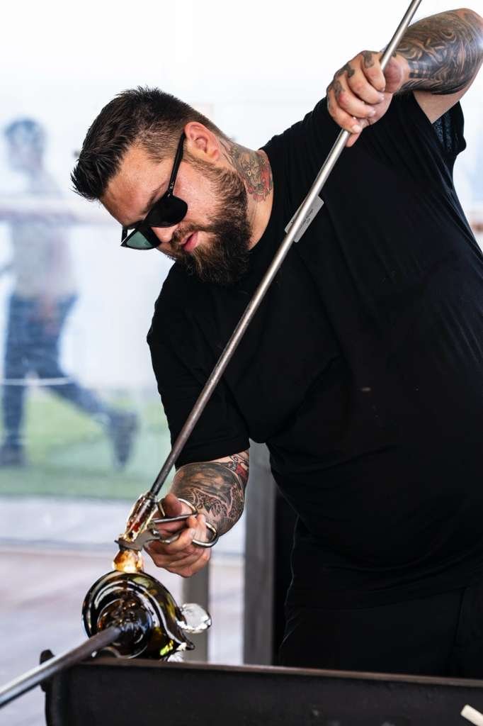 A man with tattoos and sunglasses is glassblowing, shaping molten glass on a blowpipe.