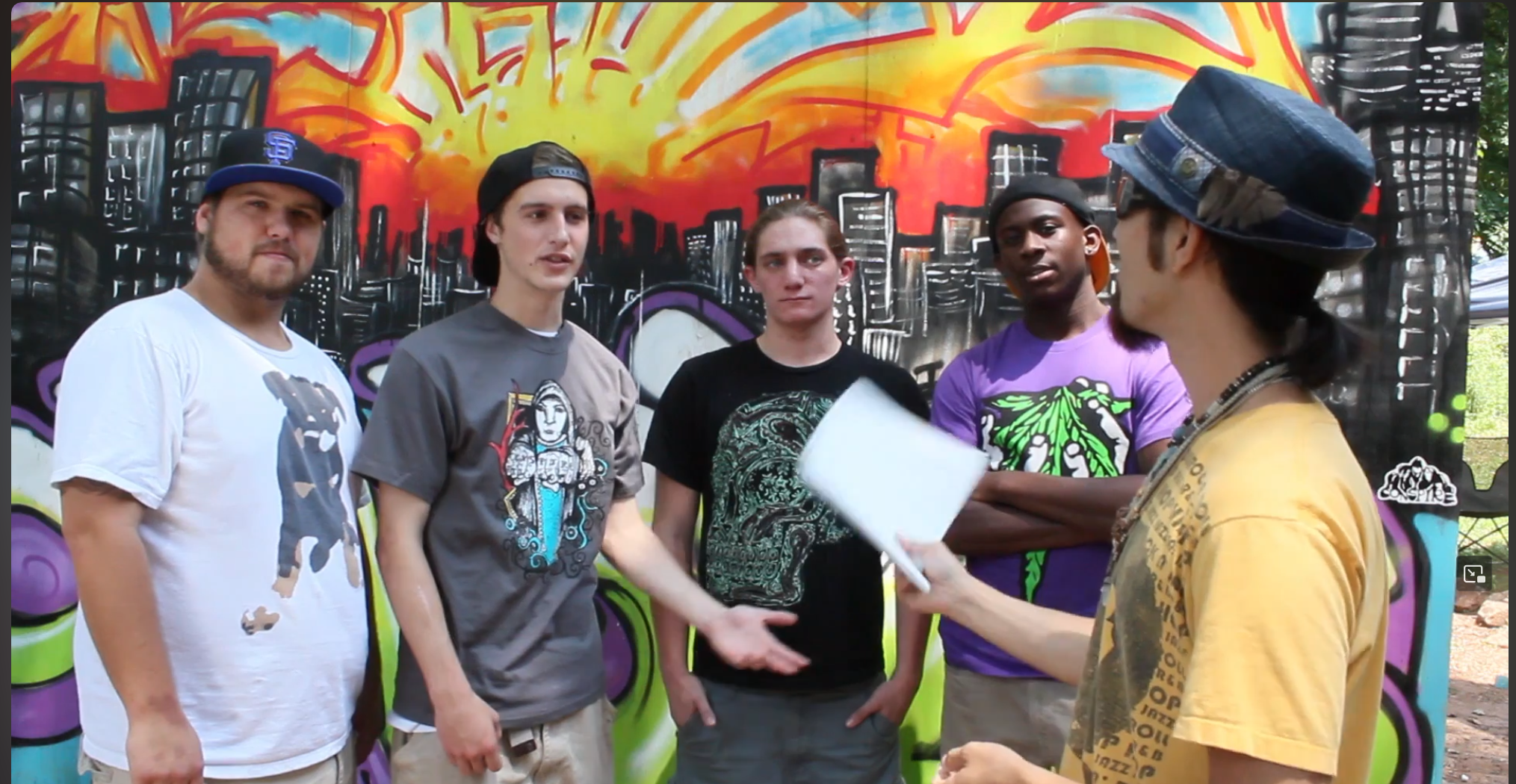 Five young men are standing in front of a vibrant graffiti wall, engaged in conversation with a woman who is holding a notebook or paper. The scene appears to be outdoors in an urban or park setting.