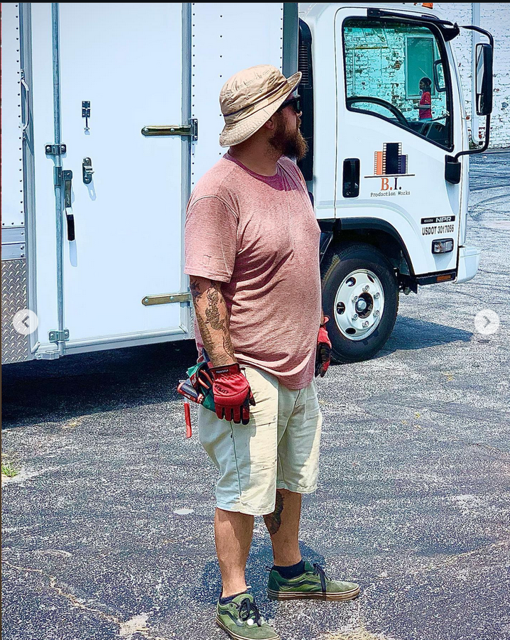A bearded man wearing a beige bucket hat, sunglasses, a pink t-shirt, and beige shorts, standing on asphalt next to a white work truck with a logo for B.I.T Production Works. He has tattoos on his left arm and is holding red work gloves in his left hand. The truck has a closed side door and a cab with window reflections showing a woman in a pink shirt.