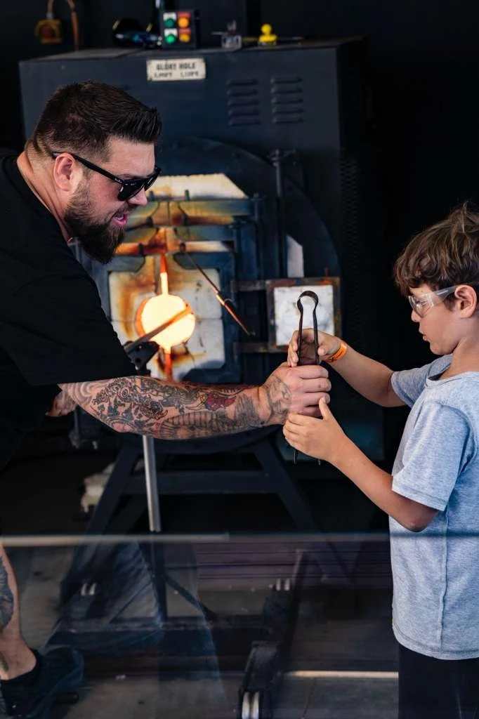 A man with tattoos on his arm and wearing sunglasses assists a young boy in holding a hot metal tool in a blacksmith's workshop. The boy wears safety glasses, and a forge is visible in the background, glowing orange with a piece of metal inside.