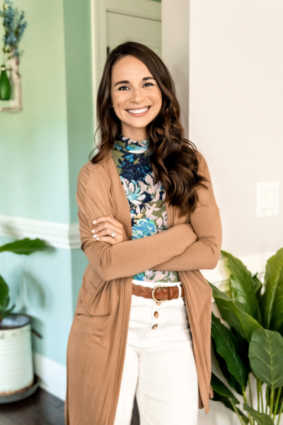 A woman with long brown hair smiling and standing indoors, wearing a floral top, beige cardigan, and white pants, with green plants and a light-colored wall in the background.