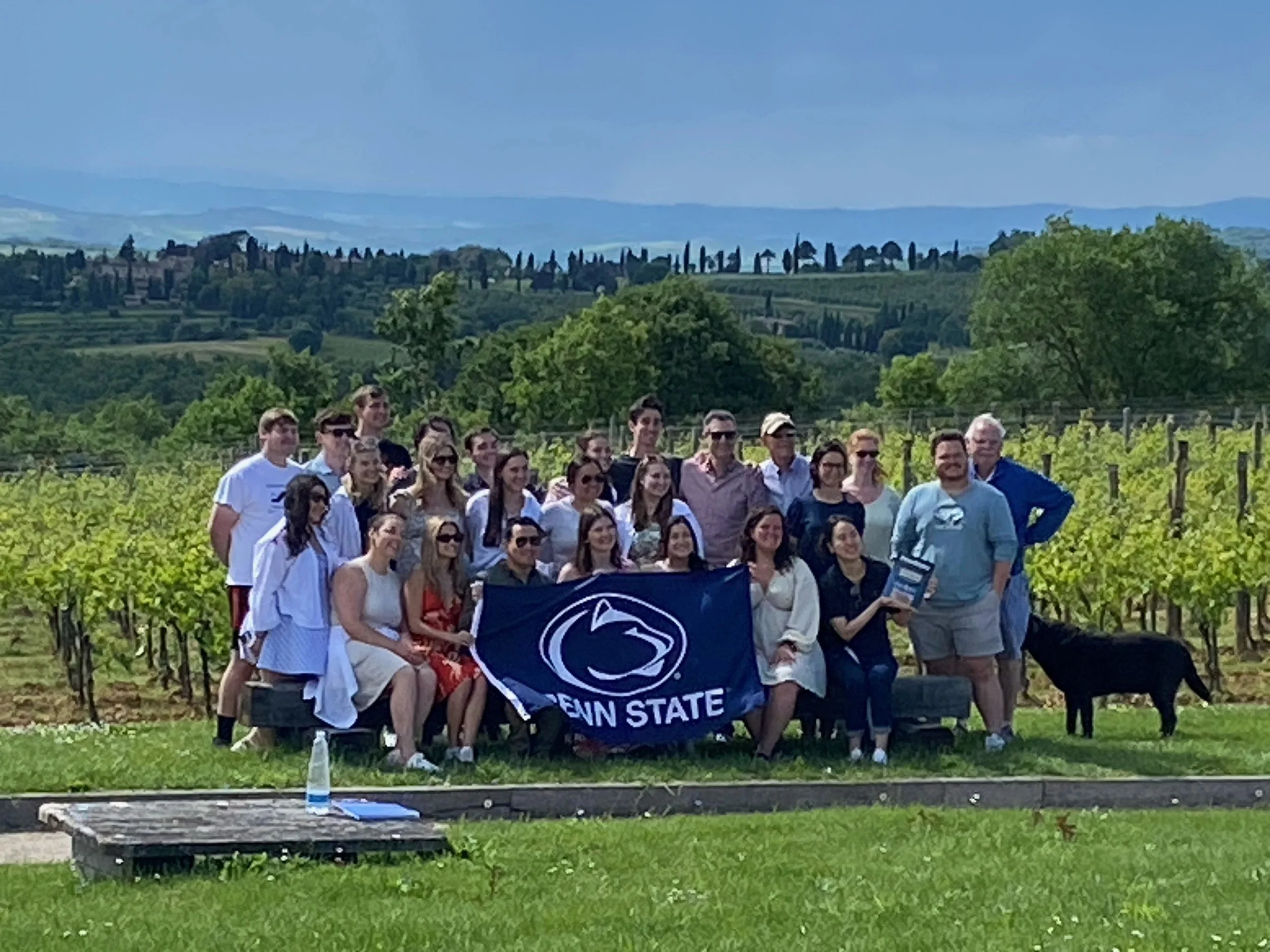 Group of people standing outdoors among vineyard rows, holding a Penn State banner, with scenic rolling hills and cypress trees in the background.