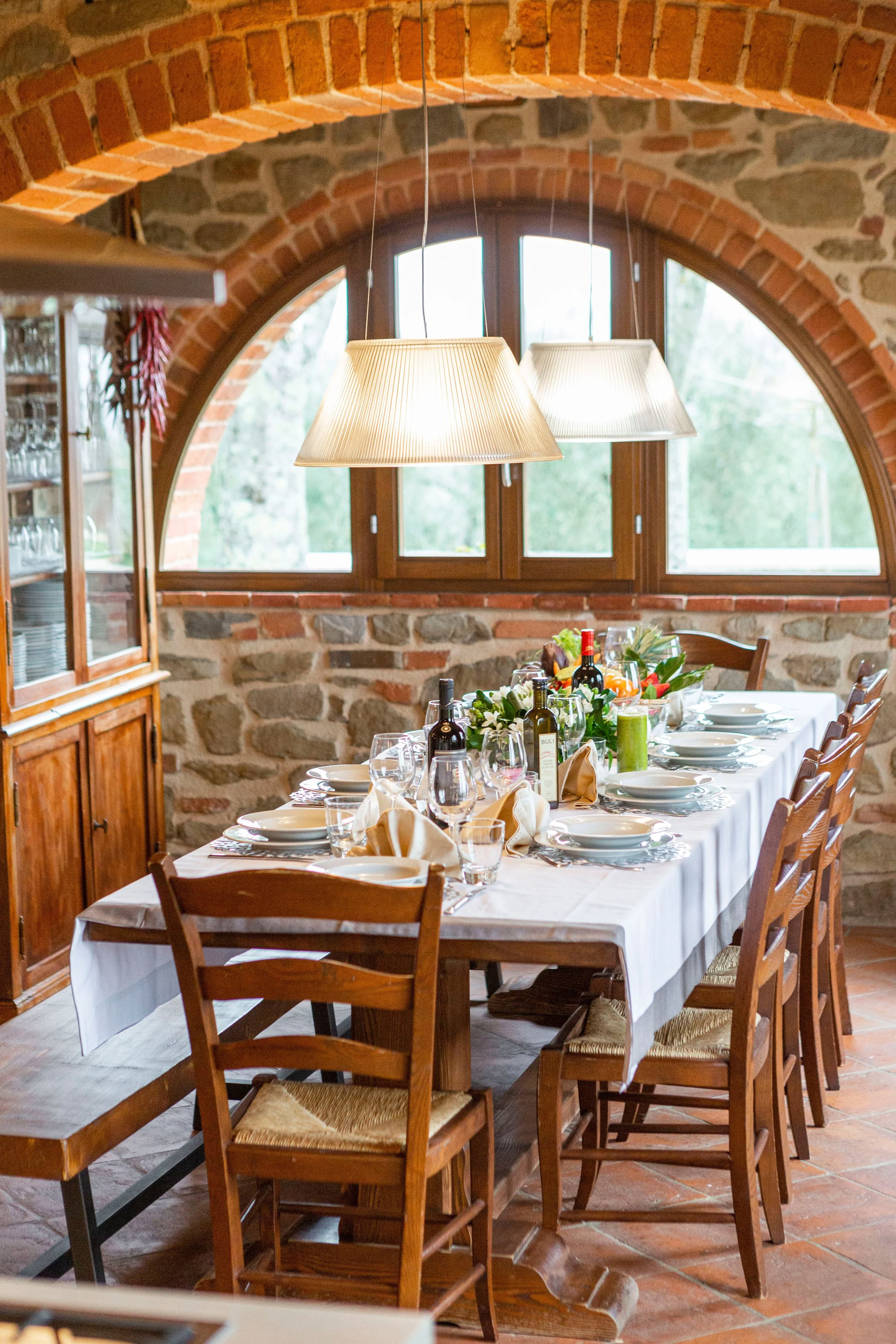A dining table set for a meal in a room with brick and stone walls, illuminated by two hanging lamps. The table has wine bottles, glasses, plates, and floral decorations.