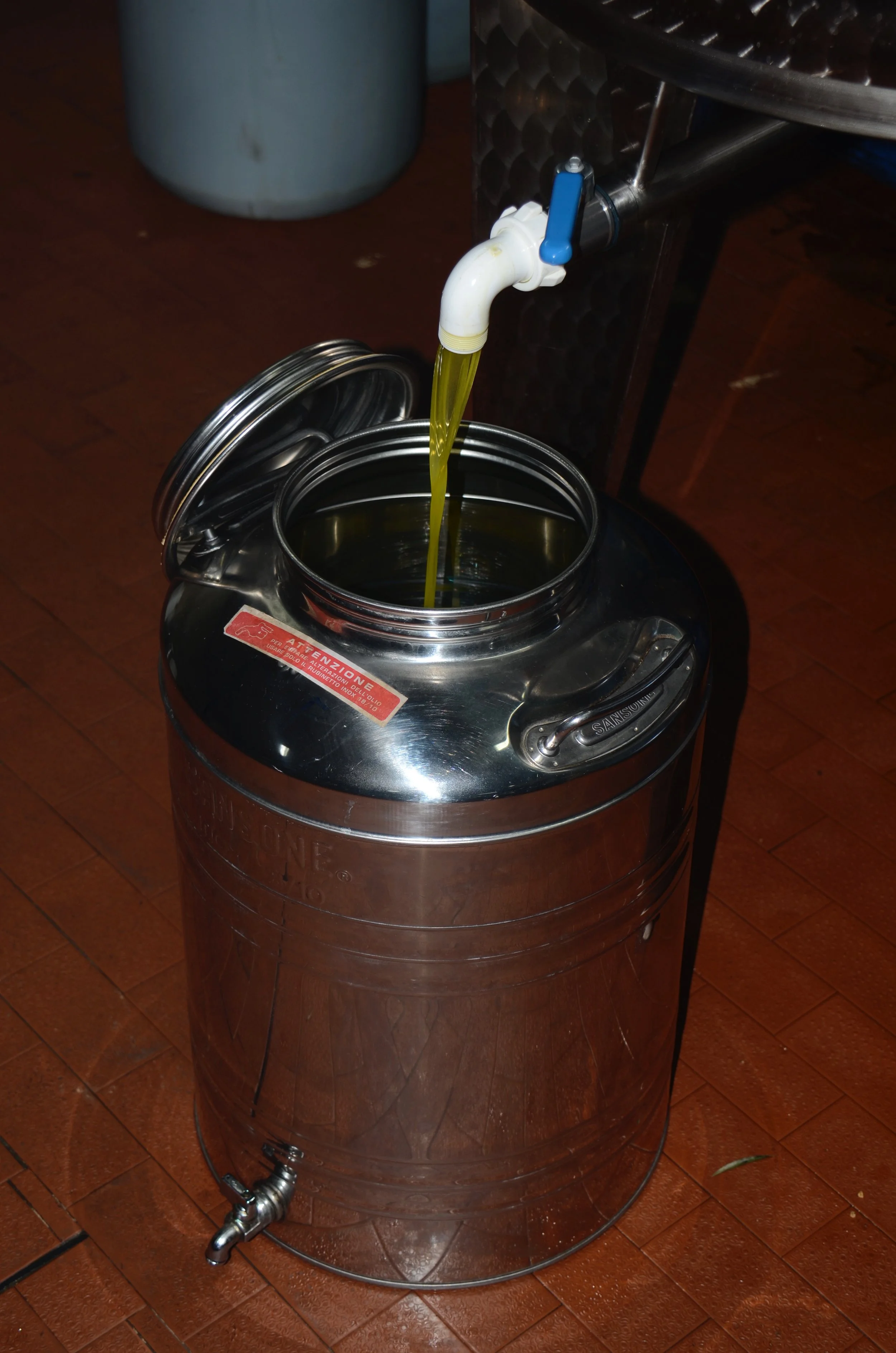 A stainless steel container with a tap, and yellow liquid flowing into it, is placed on a tiled floor.