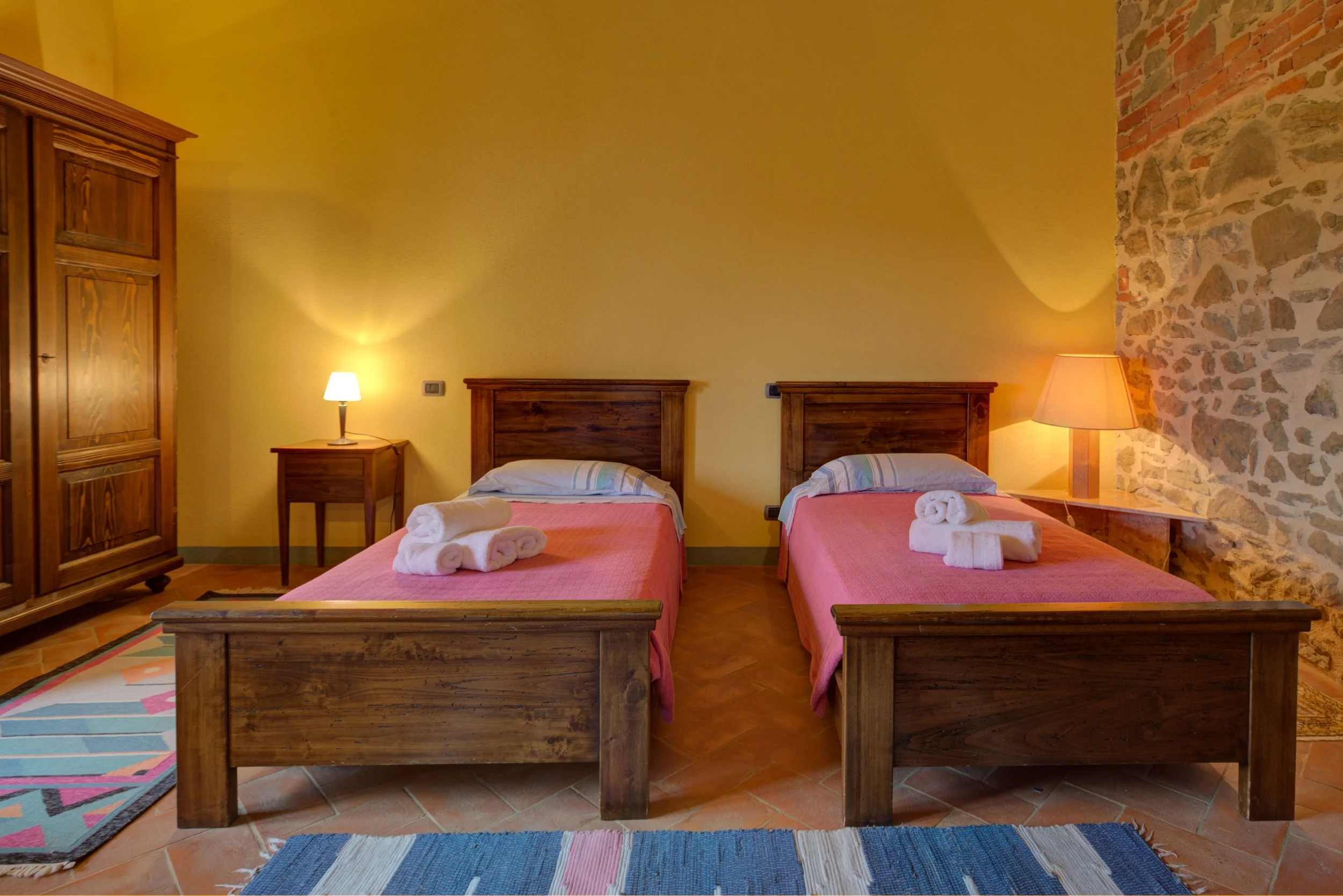 A cozy bedroom with two twin beds featuring wooden headboards and pink bedspreads. Each bed has a folded towel rolled at the foot. Between the beds are side tables with lamps, and the room has a yellow wall and stone accent wall. A wooden wardrobe is