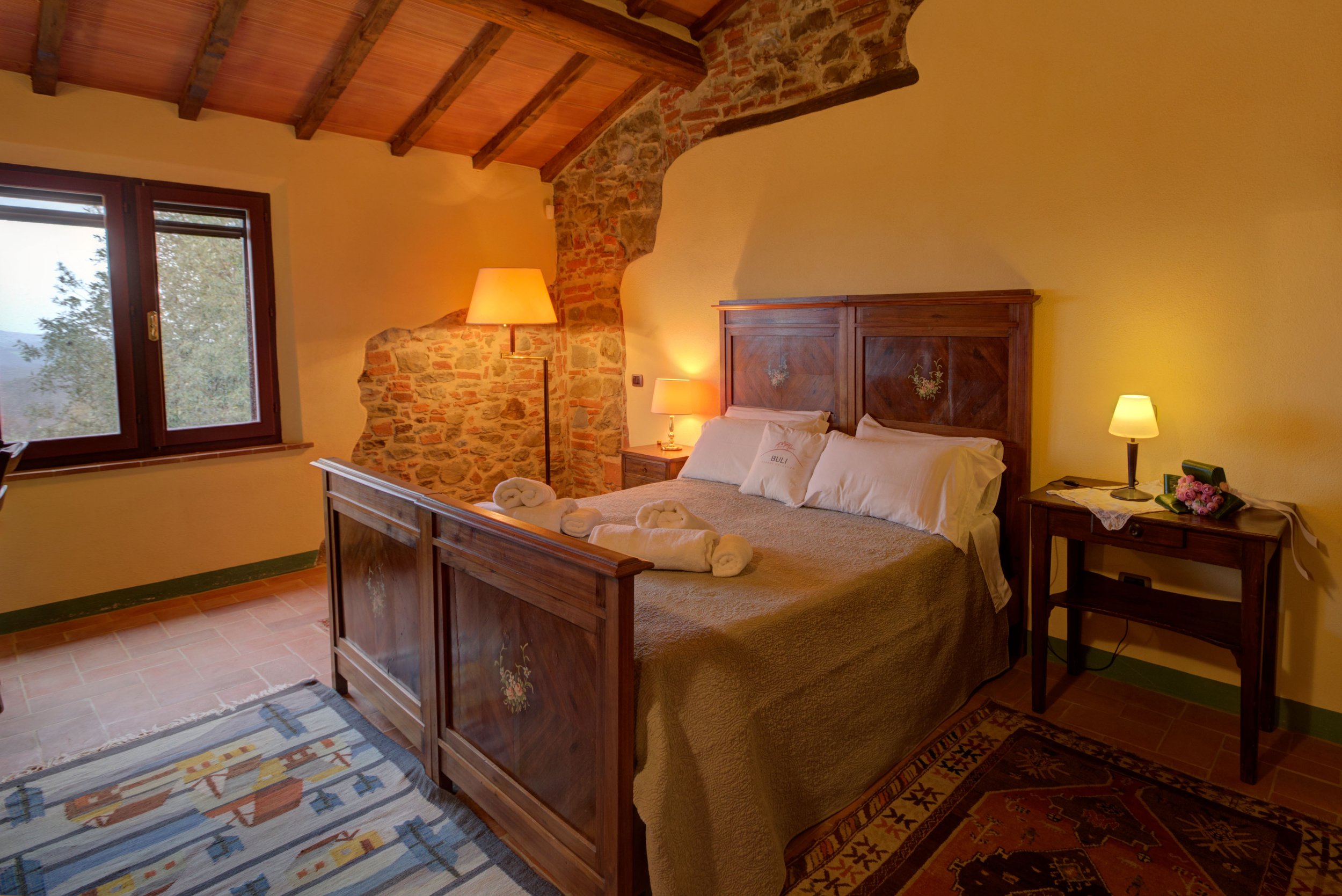Cozy bedroom with a wooden bed frame, yellow walls, and a brick and wood ceiling. The bed is made with white linens and pillowcases, with rolled towels placed on top. There are two bedside lamps, a window with a view of trees, and a colorful rug on t