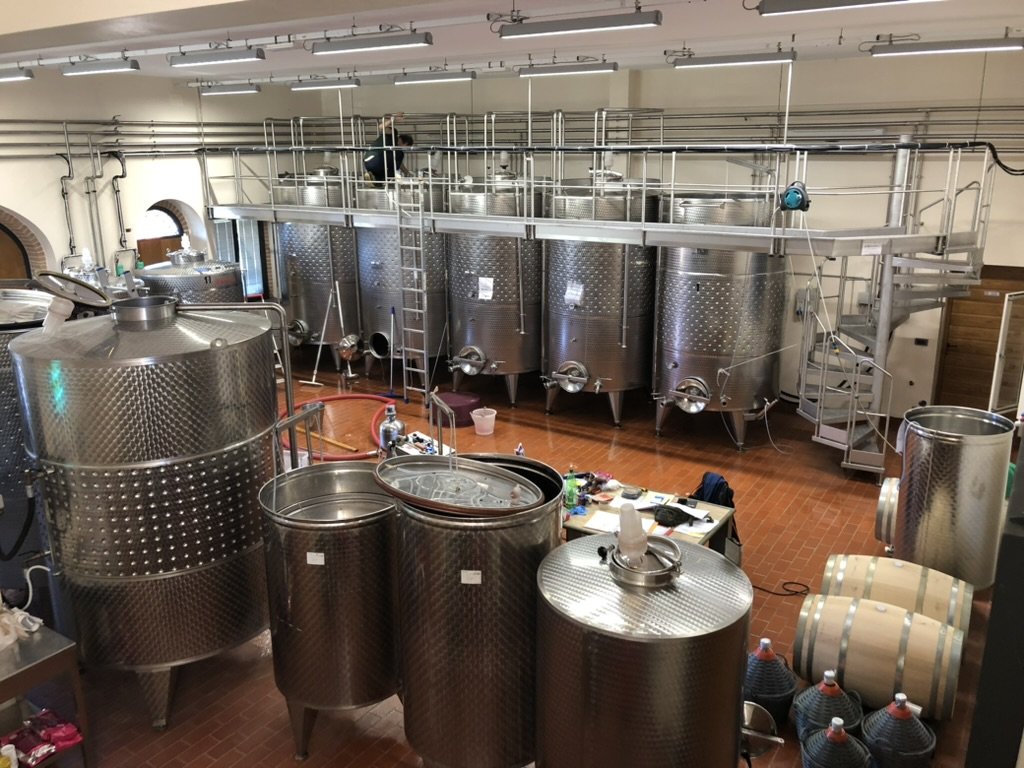 Multiple stainless steel fermentation tanks in a winery or brewery setup with a ladder and equipment around.