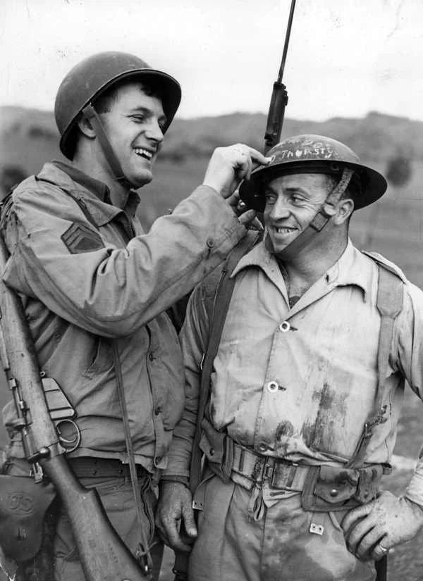 Two soldiers in uniform and helmets share a lighthearted moment, with one soldier adjusting the other's helmet.