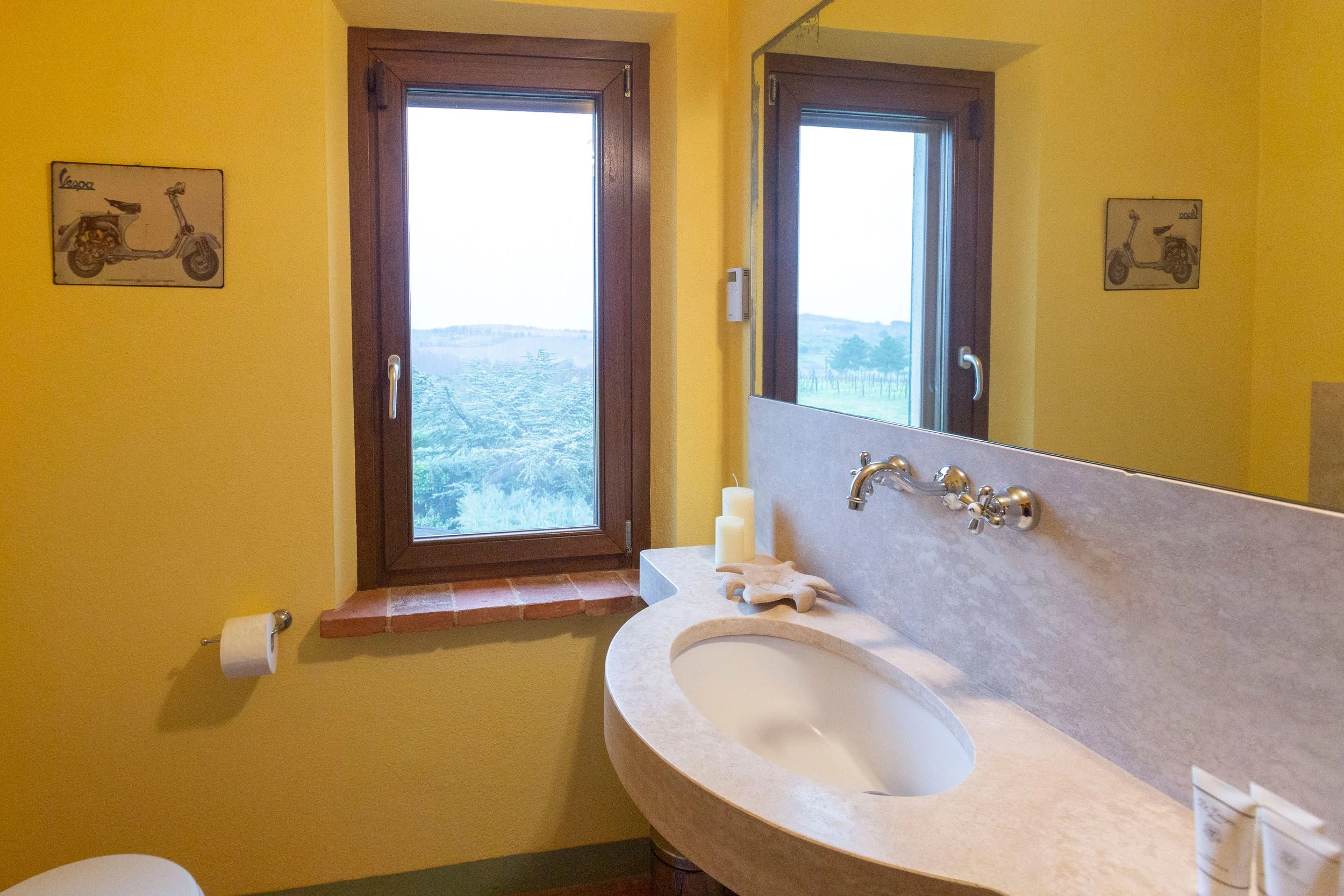 A bathroom with yellow walls. There is a large mirror above a beige curved sink with a silver faucet. Two candles in white holders, a beige towel, and a toilet paper roll are on the countertop. A window with a wooden frame shows a view of trees and h