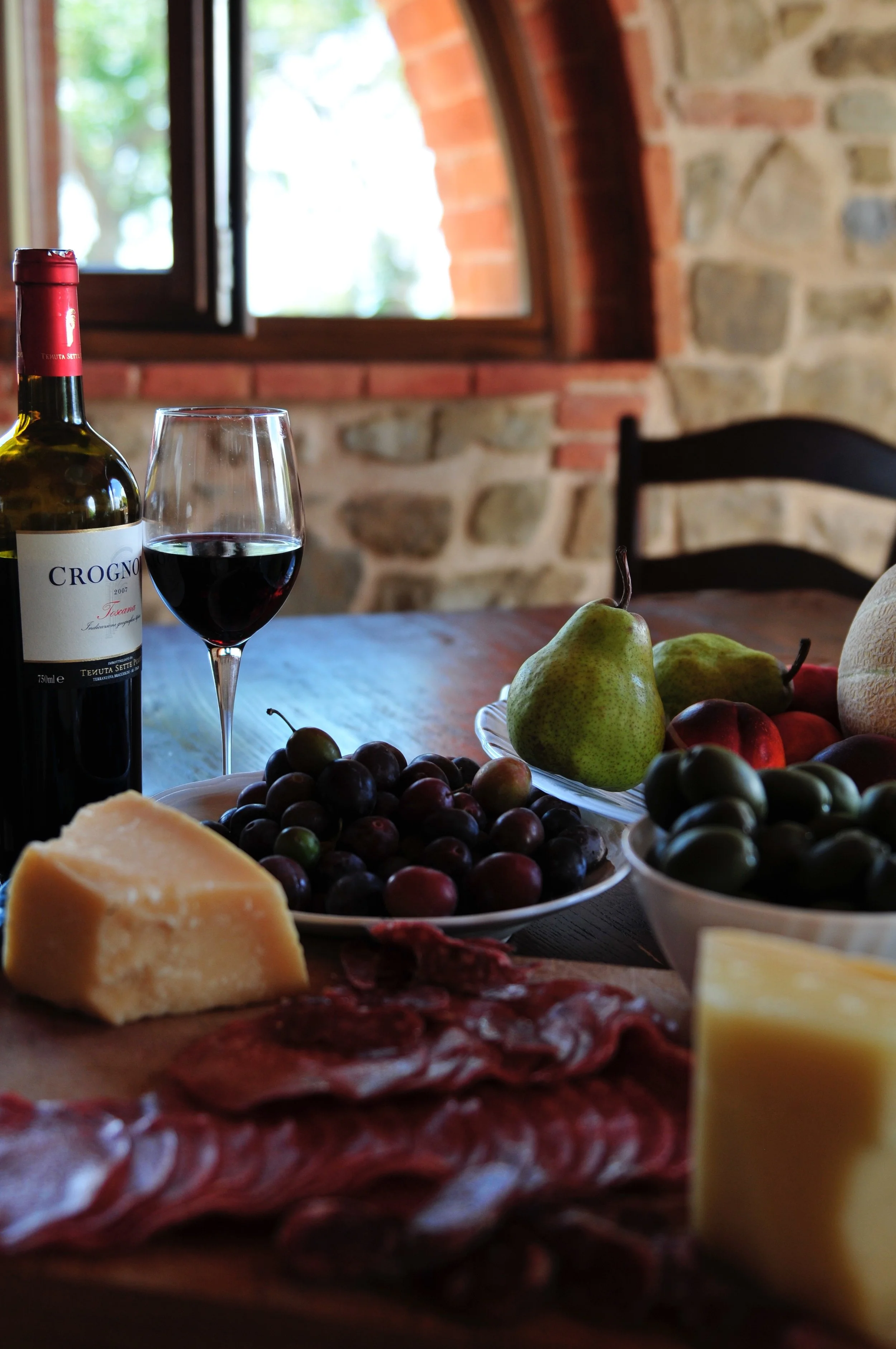 A table with a bottle of red wine, a glass of red wine, a piece of cheese, grapes, pears, apples, and other assorted food items in a rustic room with a brick and stone wall and an open window.