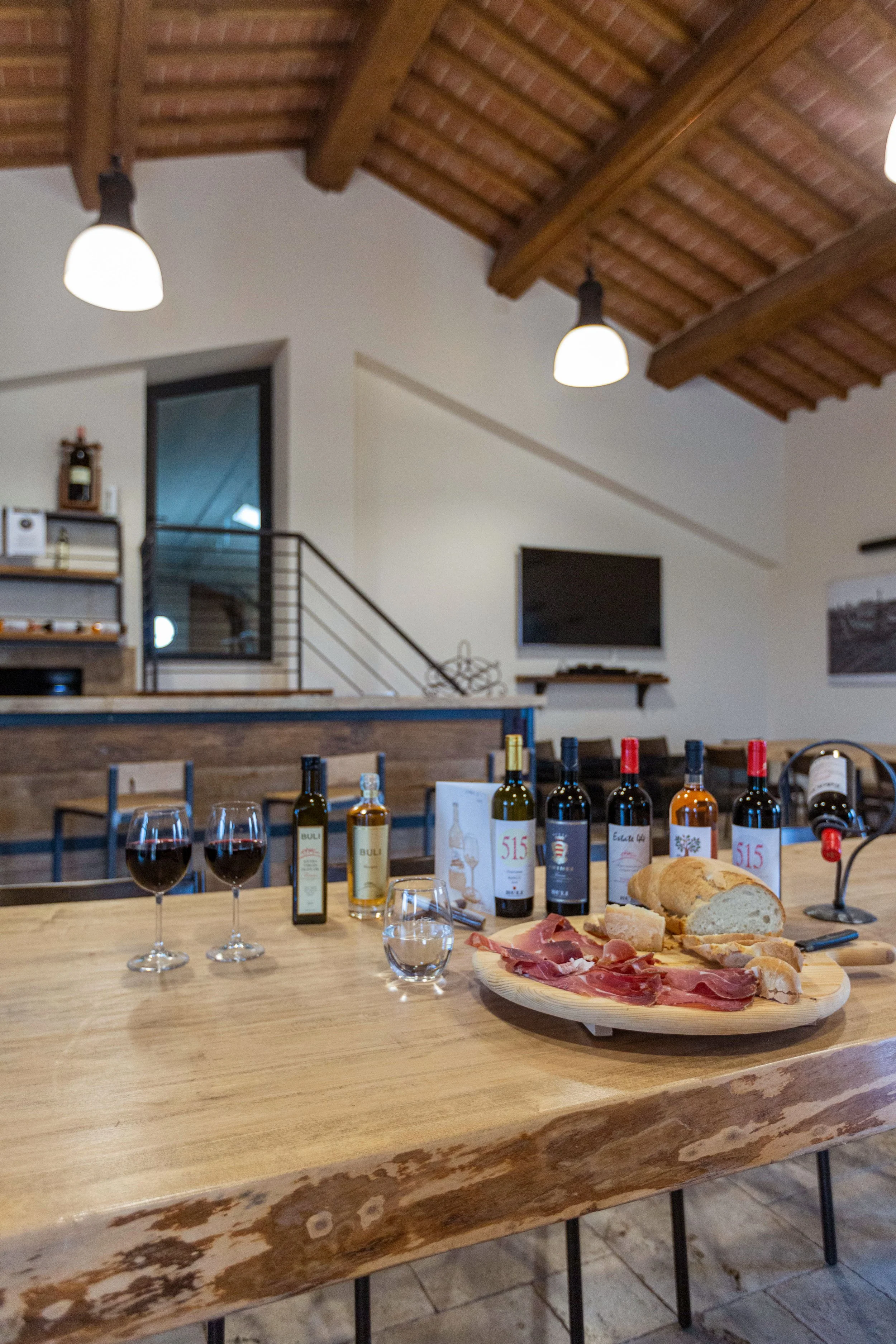 A wooden table set with bottles of wine, glasses of red wine, and a cheese and charcuterie platter, in a cozy, rustic kitchen with wooden beams and modern lighting.