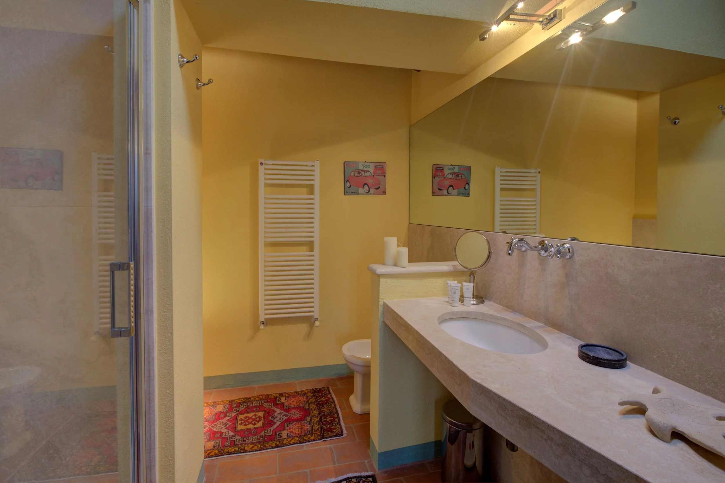 A bathroom with yellow walls, a large mirror, a marble sink countertop, a small vanity mirror, and wall-mounted towel heaters. Two candles are on the counter and a colorful patterned rug on the tile floor.