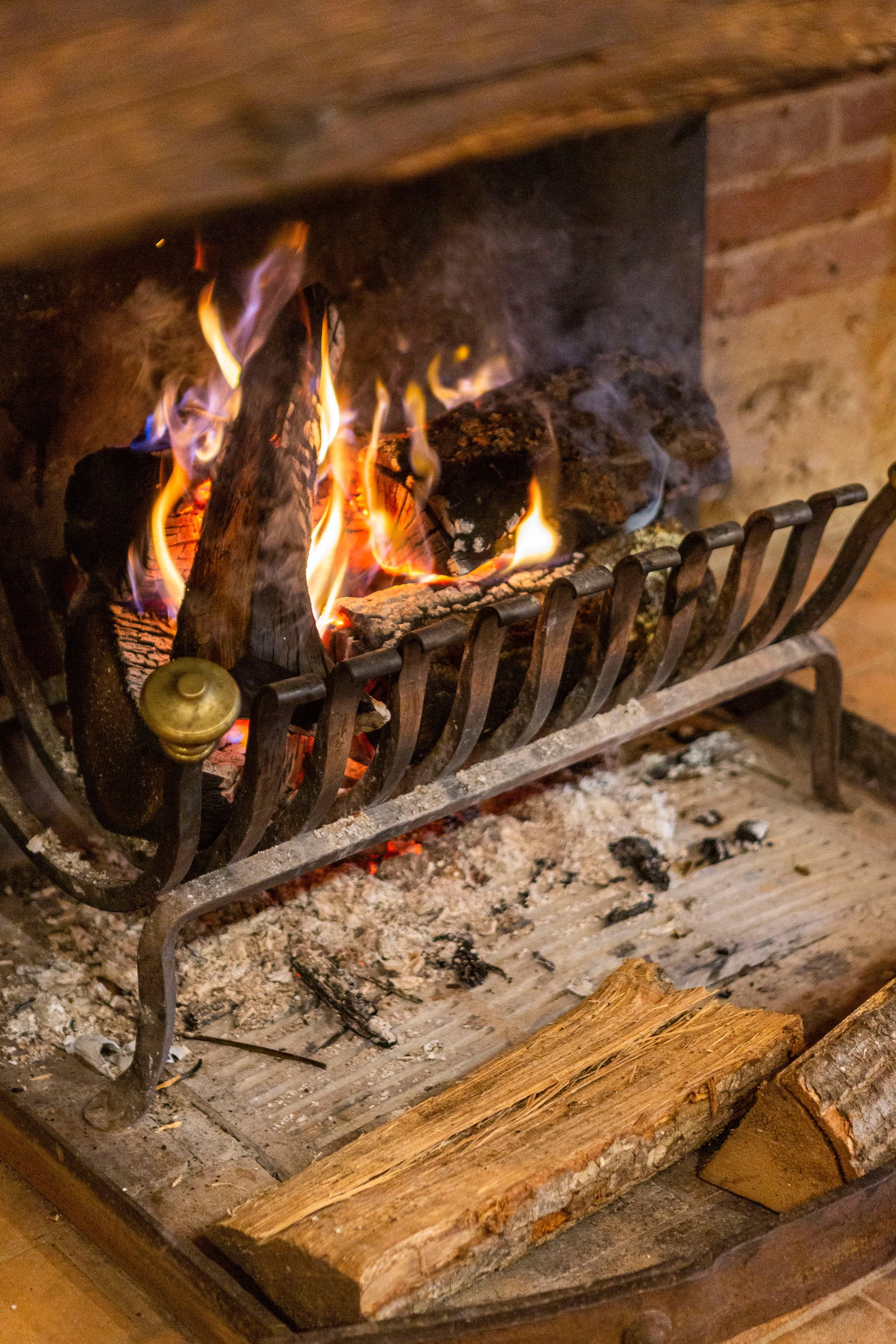 A close-up of a fireplace with burning logs and flames, showing a mix of firewood, ashes, and a metal grate.