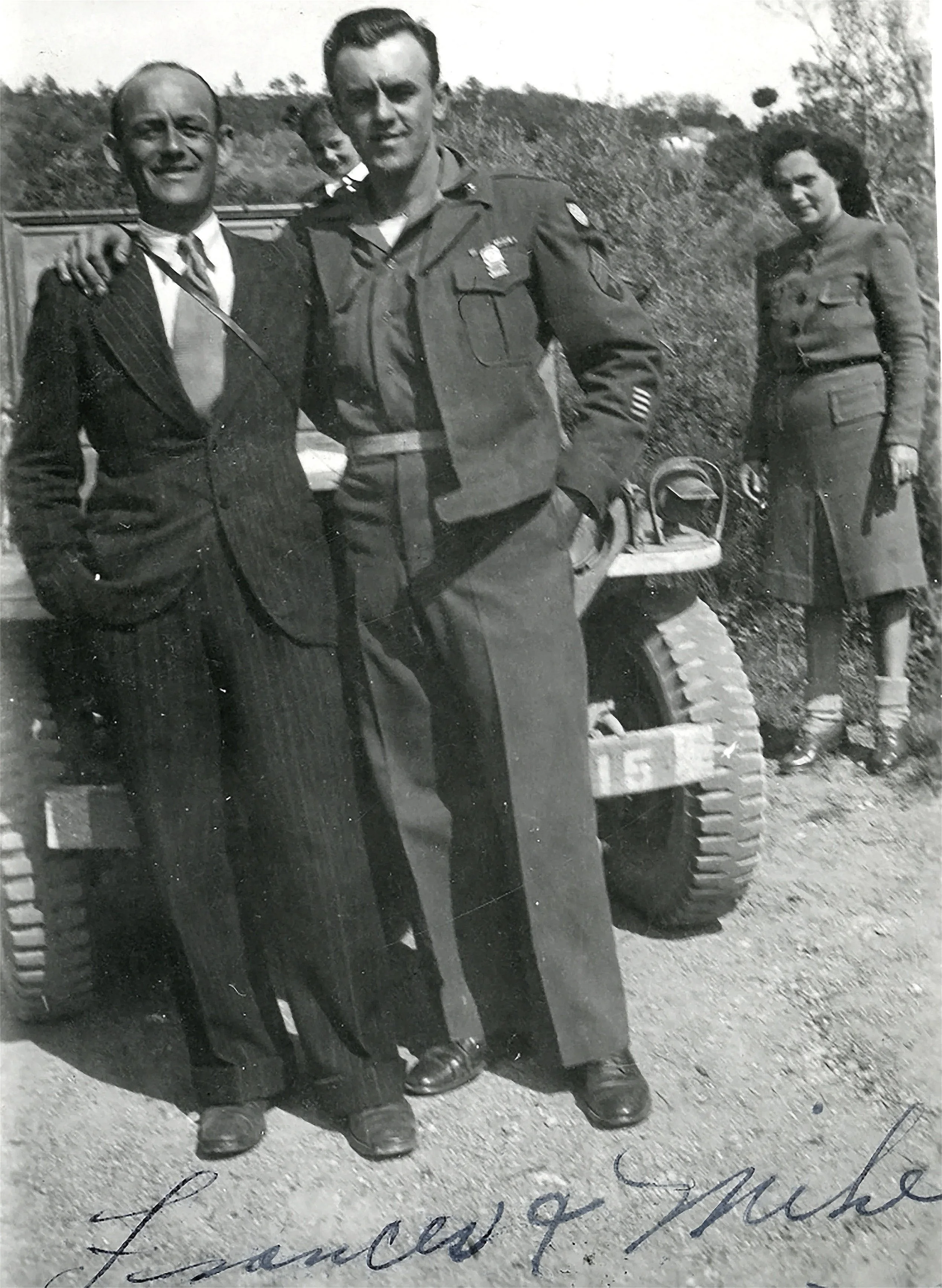 Black and white photograph of a group of people outdoors, including a man in military uniform, a man in a suit, a woman, and a young girl, standing beside a military vehicle with large tires.