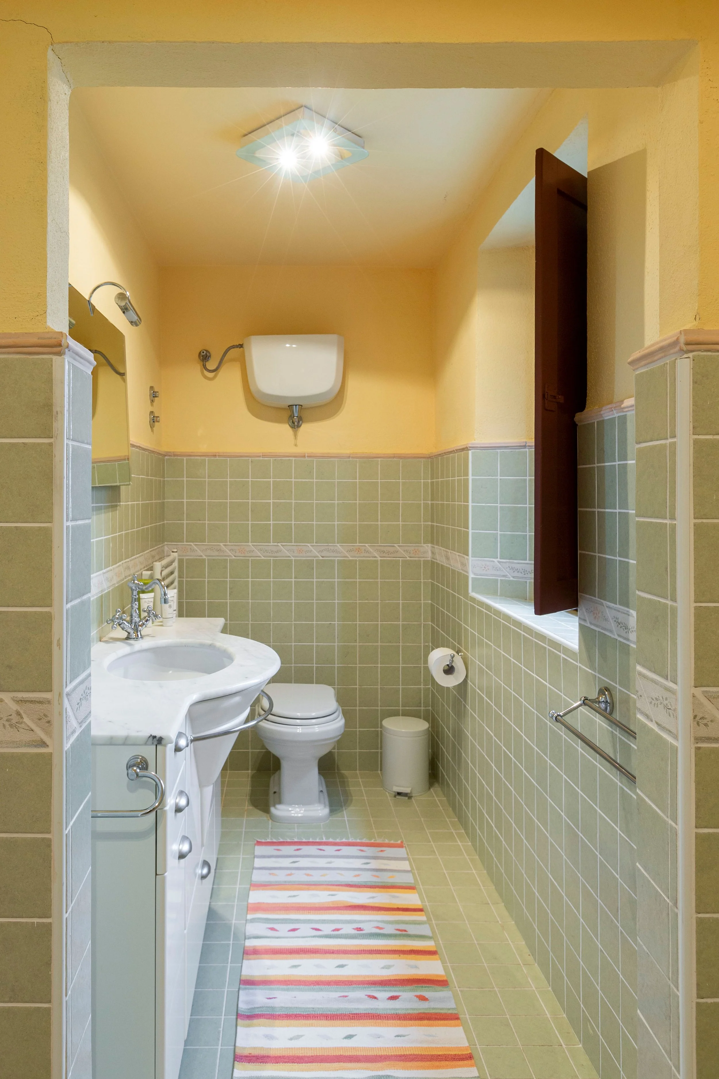 A small bathroom with yellow walls and green tiles, featuring a toilet, a small white sink, a mirror, a window with a dark wooden shutter, and a colorful striped rug on the tiled floor.