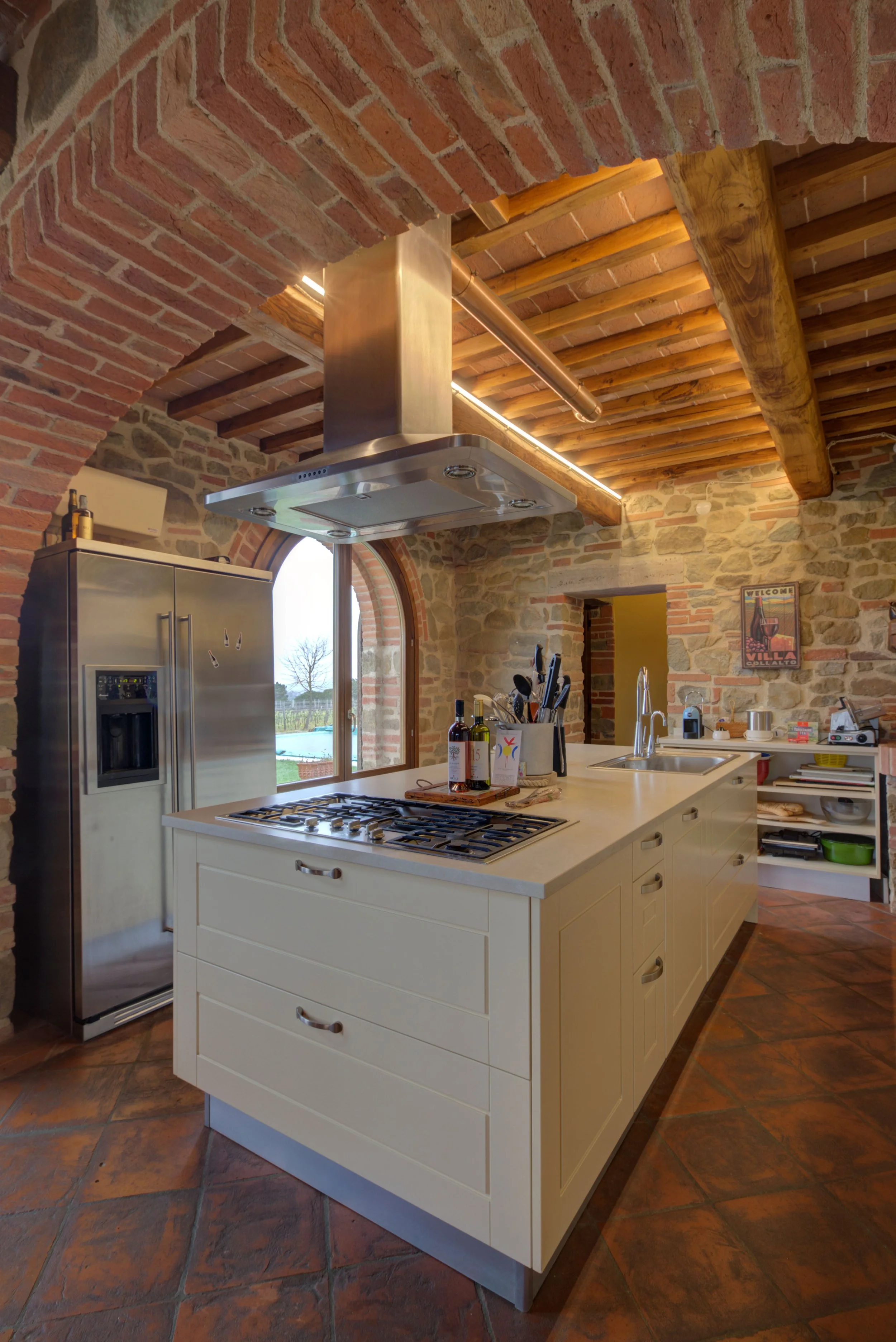 Kitchen with exposed brick walls, a kitchen island, and stainless steel appliances, including a refrigerator. The ceiling has wooden beams and brick arch additions, with natural light coming through a large arched window.