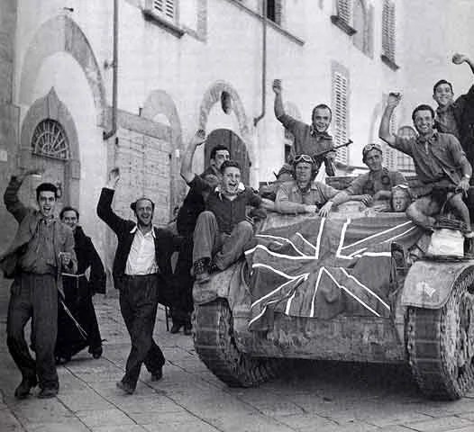 Group of smiling soldiers celebrating on and around a tank with a Union Jack flag in an urban street.