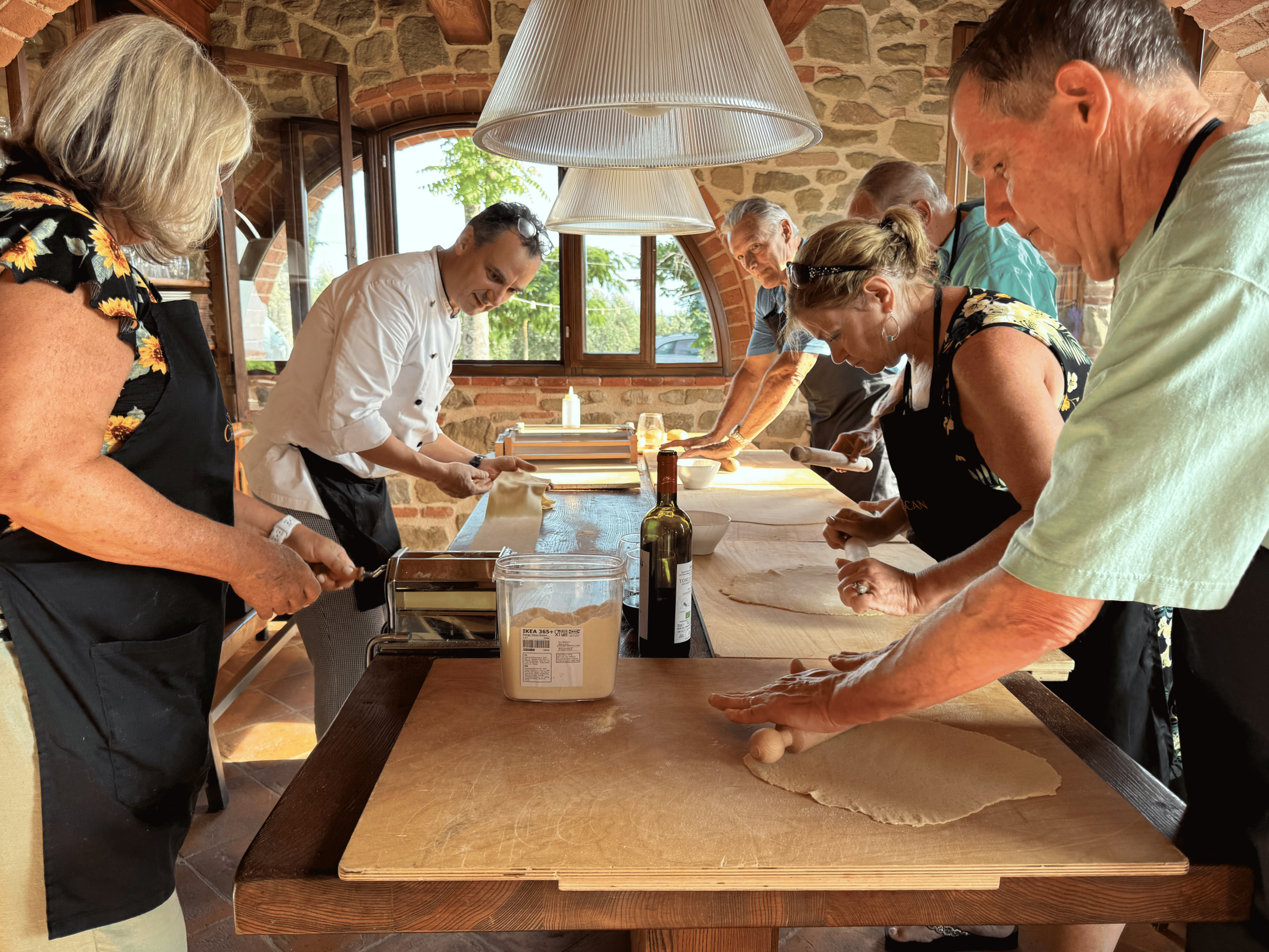 People gathered around a wooden table preparing food in a rustic kitchen with stone walls and large windows, overseen by a chef in white.