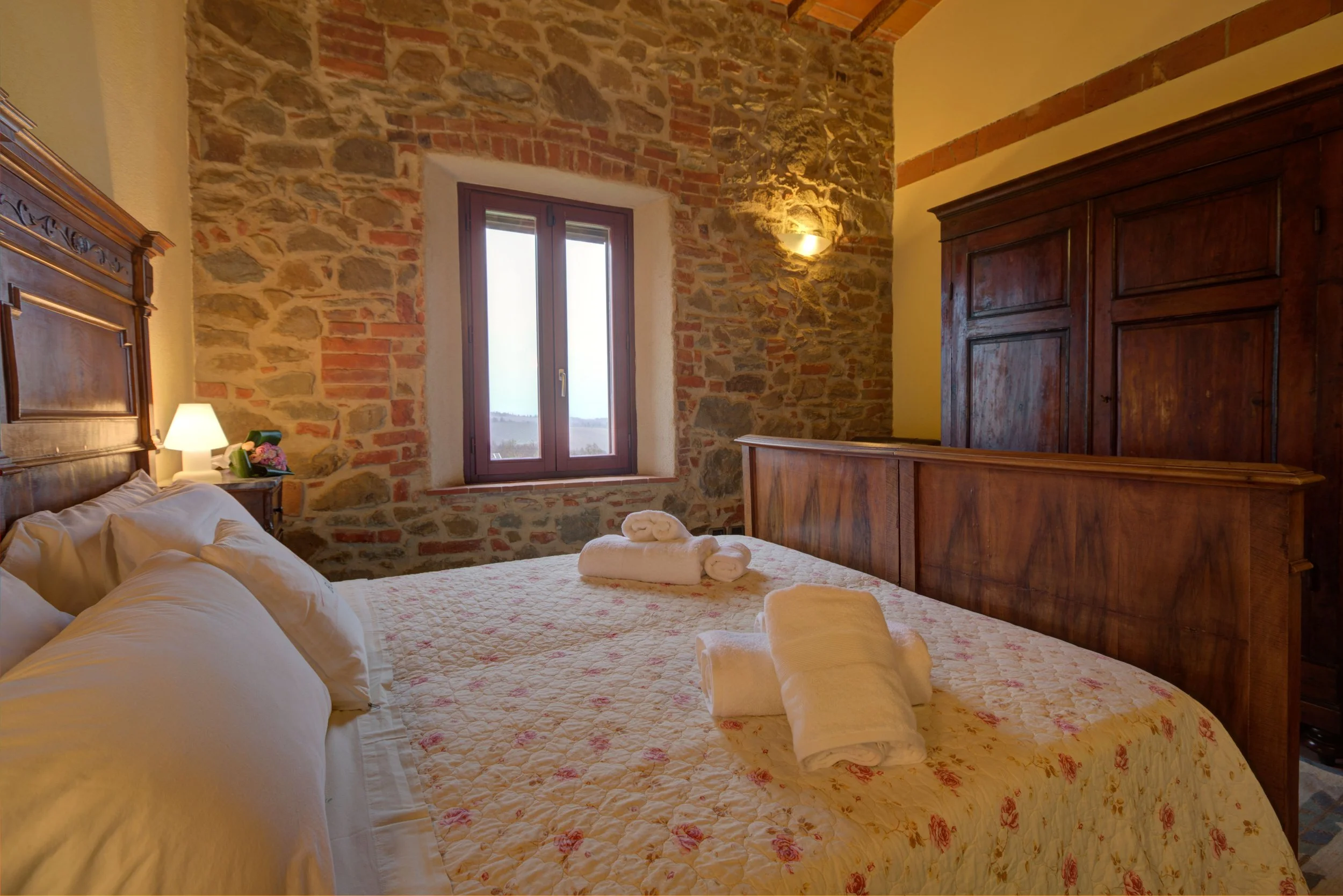A cozy bedroom with a stone wall, a window, a wooden wardrobe, a bed with white bedding and folded towels, a small bedside table with a lamp, and a floral quilt.