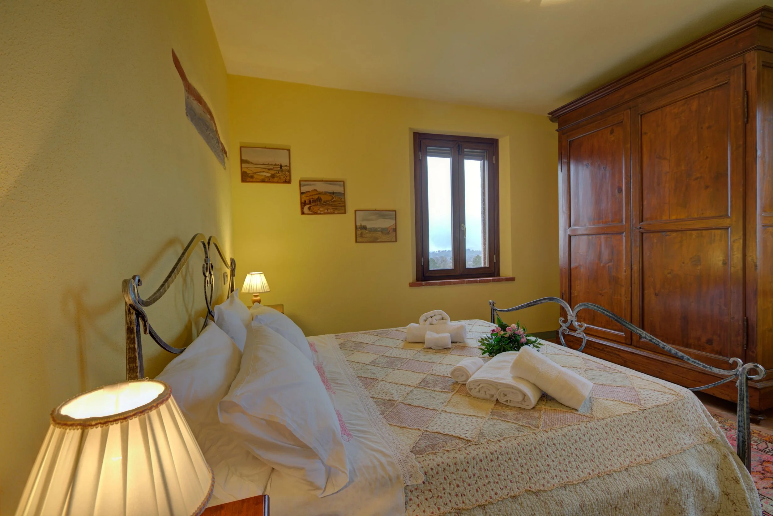 A cozy bedroom with yellow walls and a wooden wardrobe, featuring a bed with white pillows and bedspread, a bedside lamp, and window with a view of the outdoors.