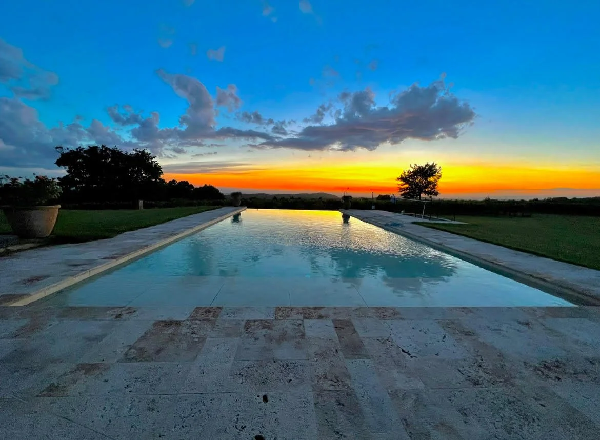 An outdoor swimming pool with a sunset sky in the background, trees on the horizon, and a clear reflection of the sky on the pool's water.