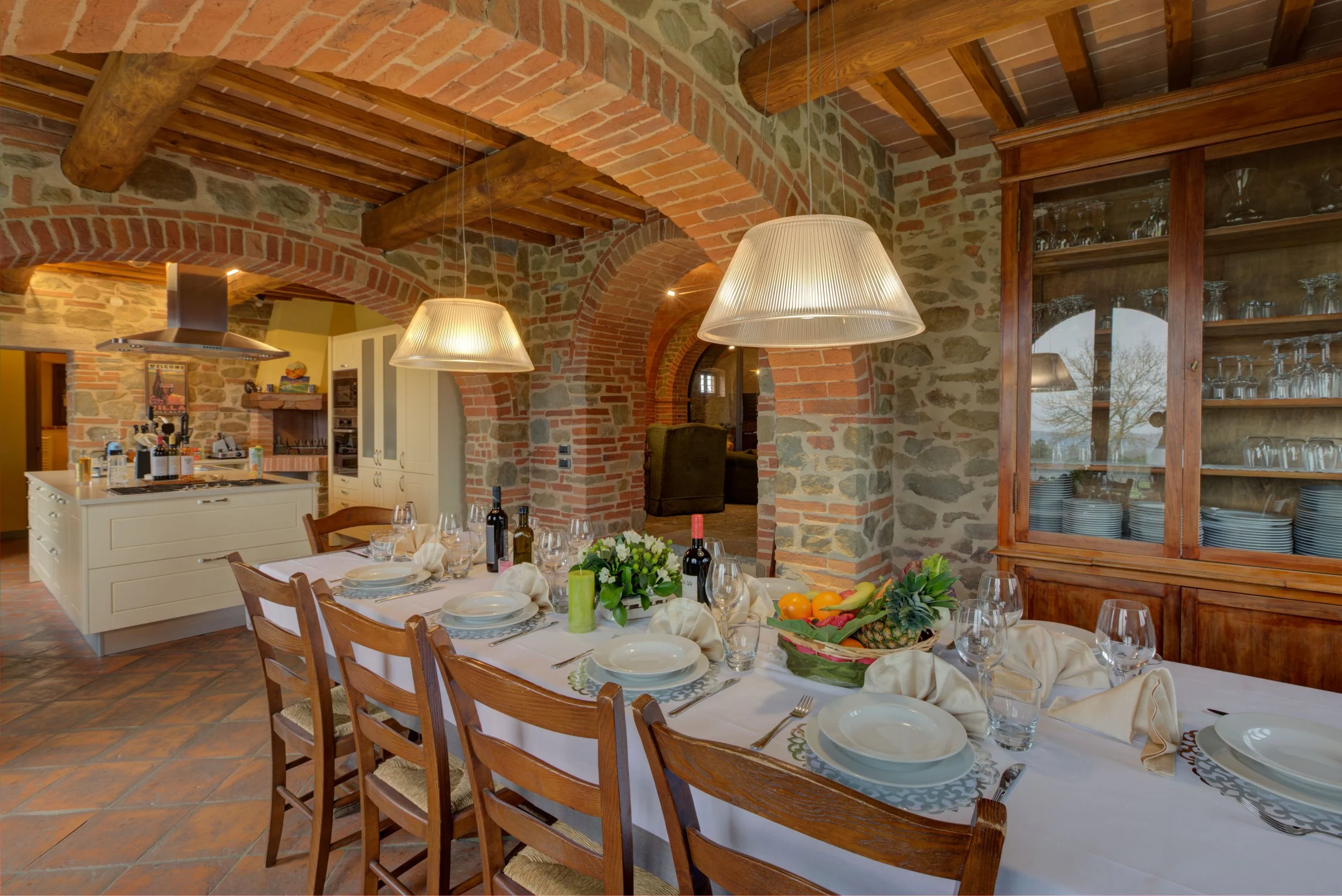 Dining room with a table set for a meal, featuring white plates, wine glasses, and utensils, surrounded by wooden chairs. The room has brick and stone walls with arched openings and exposed wooden beams on the ceiling. A sideboard with glassware is v