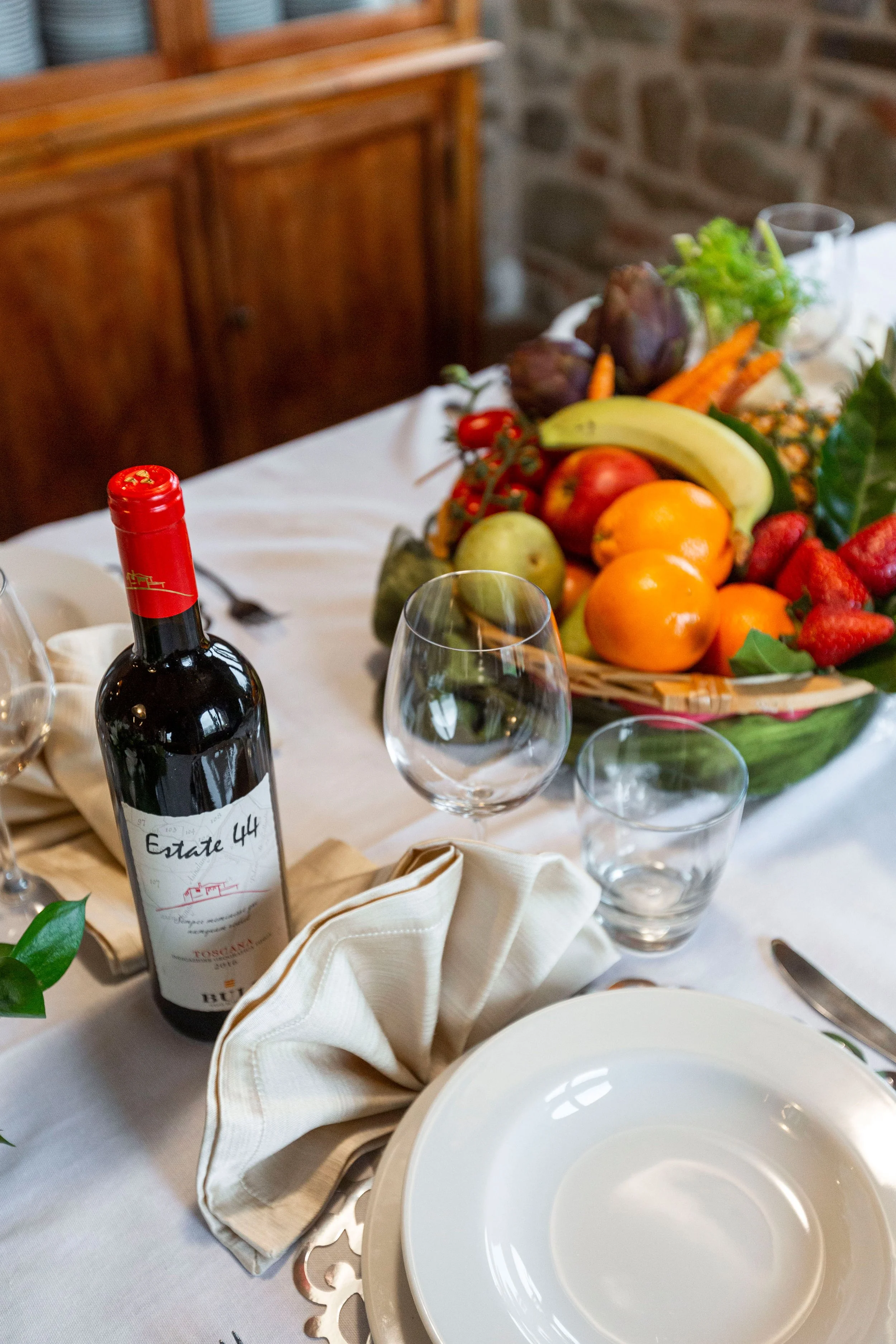 A table setting with a bottle of red wine, empty white plates, multiple glasses, a napkin, and a centerpiece of fresh fruit including bananas, oranges, strawberries, and figs, on a white tablecloth.