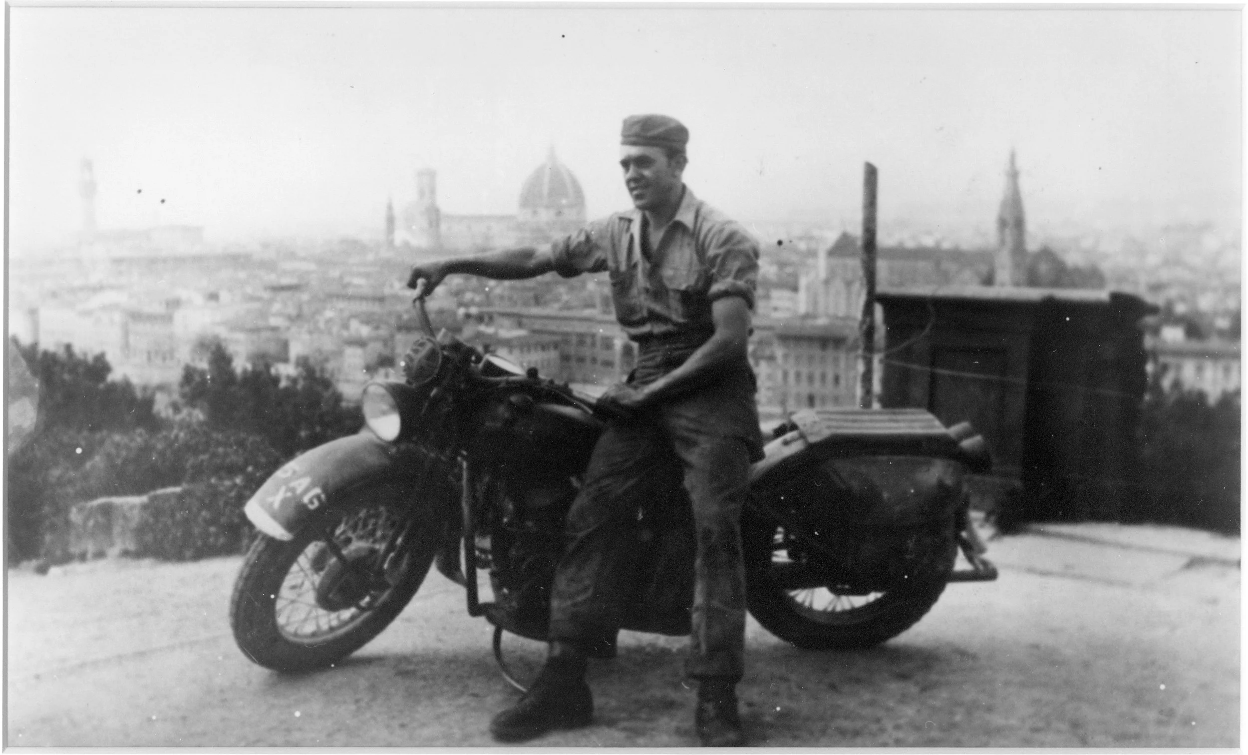 A black and white photo of a young man in military uniform sitting on a motorcycle with a sidecar, with a cityscape featuring a dome building in the background.