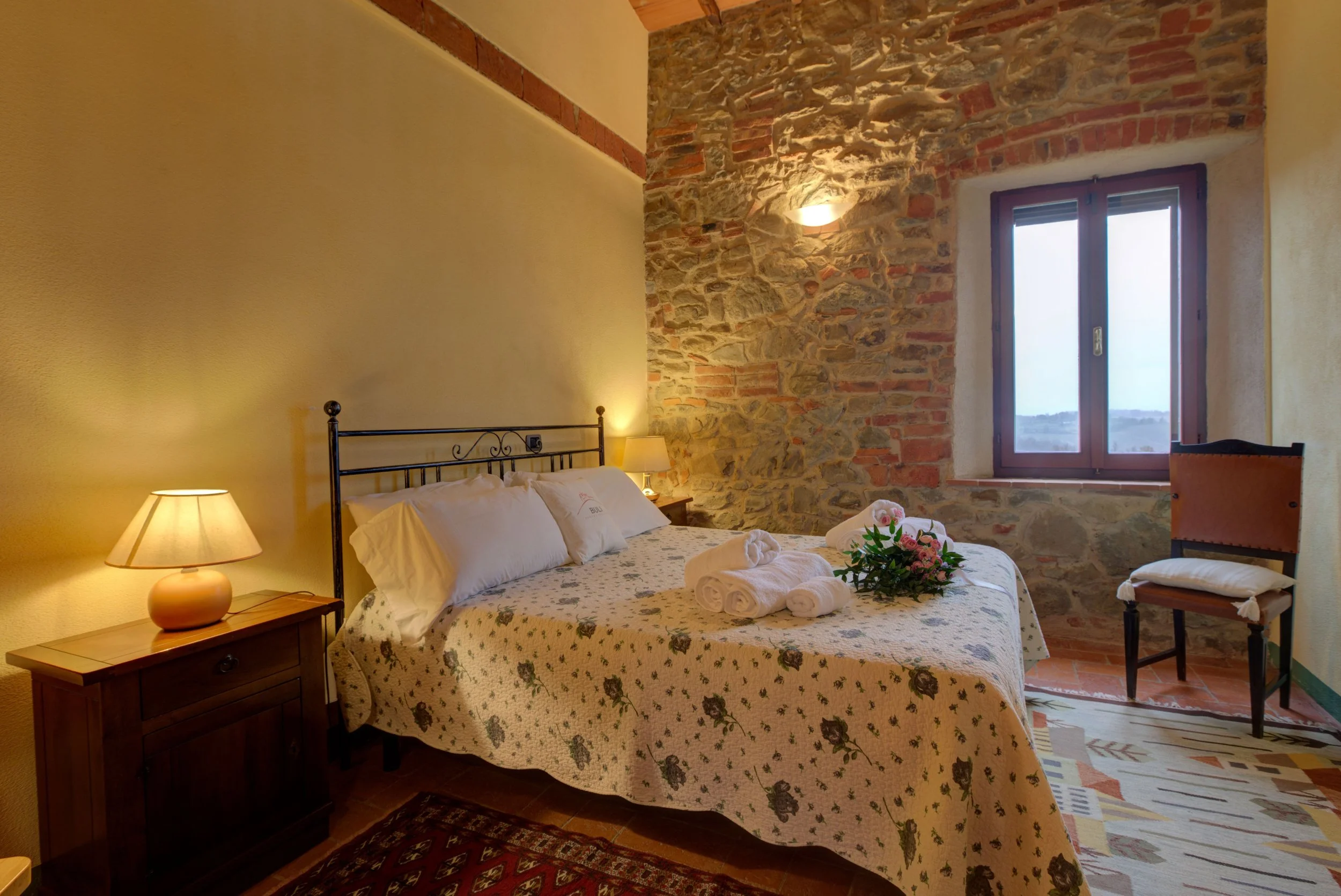 A cozy bedroom with a bed covered in a floral quilt, a nightstand with a small lamp, a vase of pink flowers, and neatly folded towels. There is a stone and brick wall behind the bed with a window that reveals a view of the outdoors.