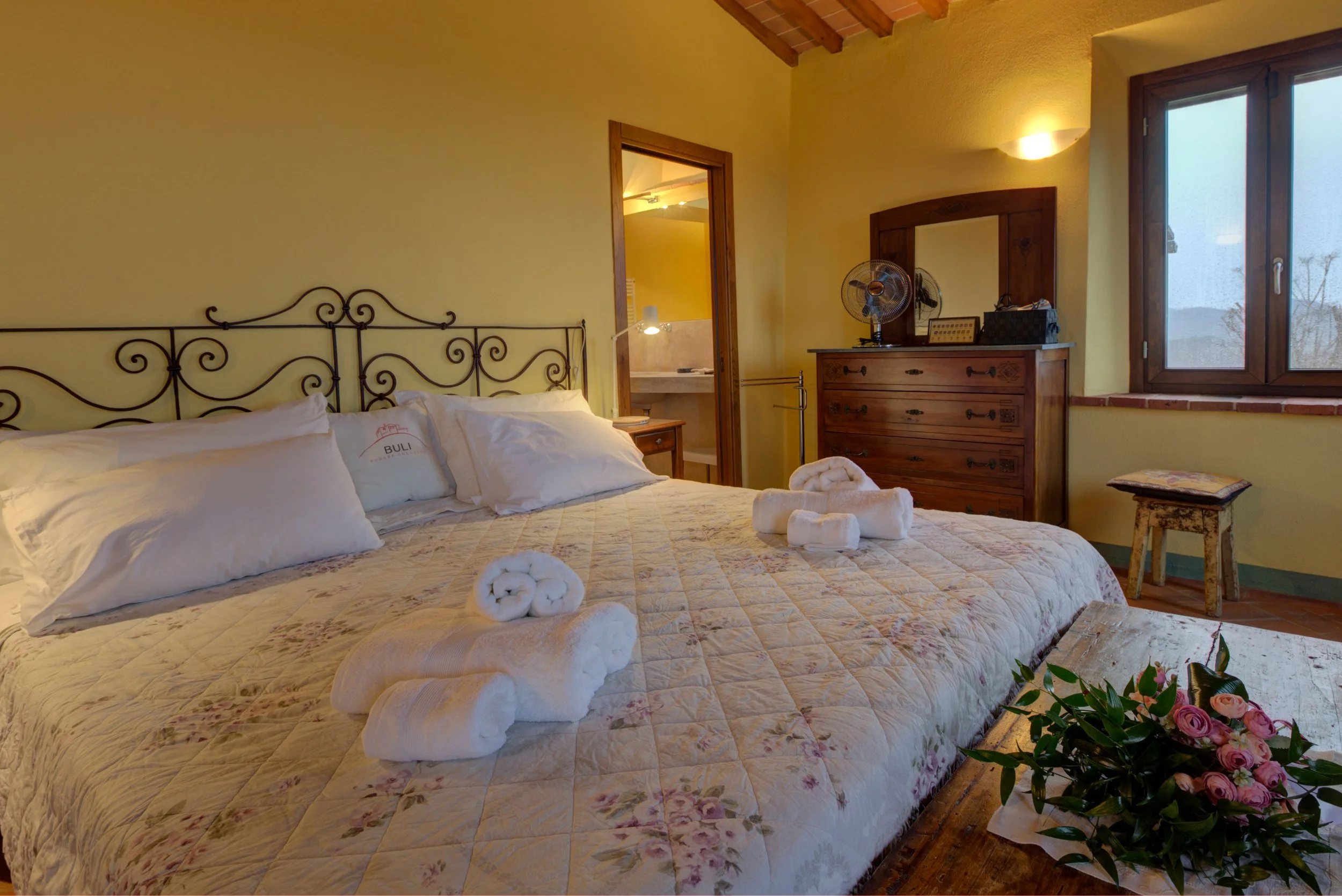A cozy bedroom with a bed covered in a floral quilt and white pillows, topped with rolled towels. A wooden dresser with a mirror and two fans is near a window, with a small table holding a floral arrangement beside the bed. The room has warm yellow w