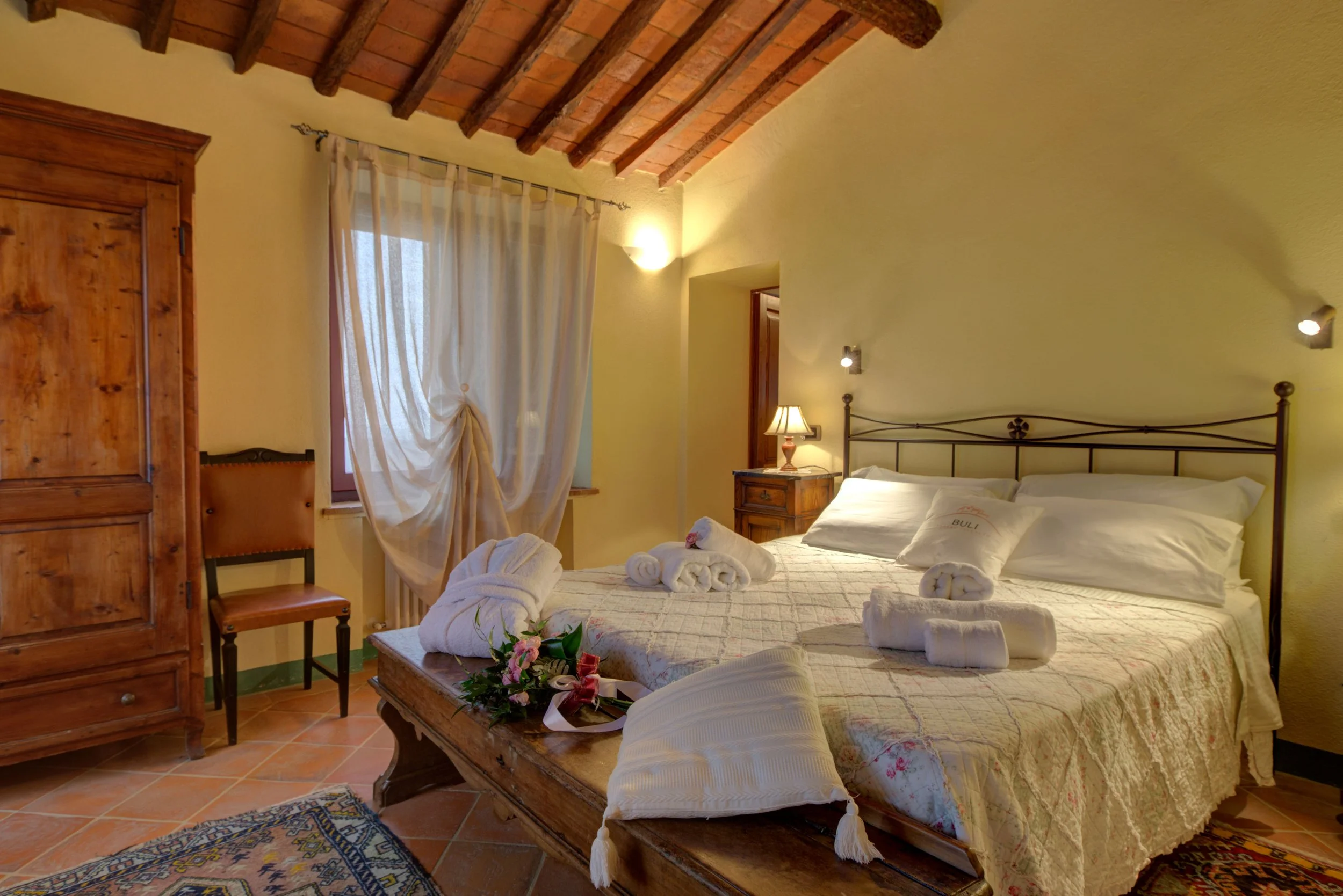 A cozy bedroom with a sloped brick ceiling, yellow walls, and wooden furniture. The bed has white linens, towels, and decorative flowers on it. There is a window with sheer curtains, a wooden wardrobe, a chair, a small bedside table with a lamp, and 