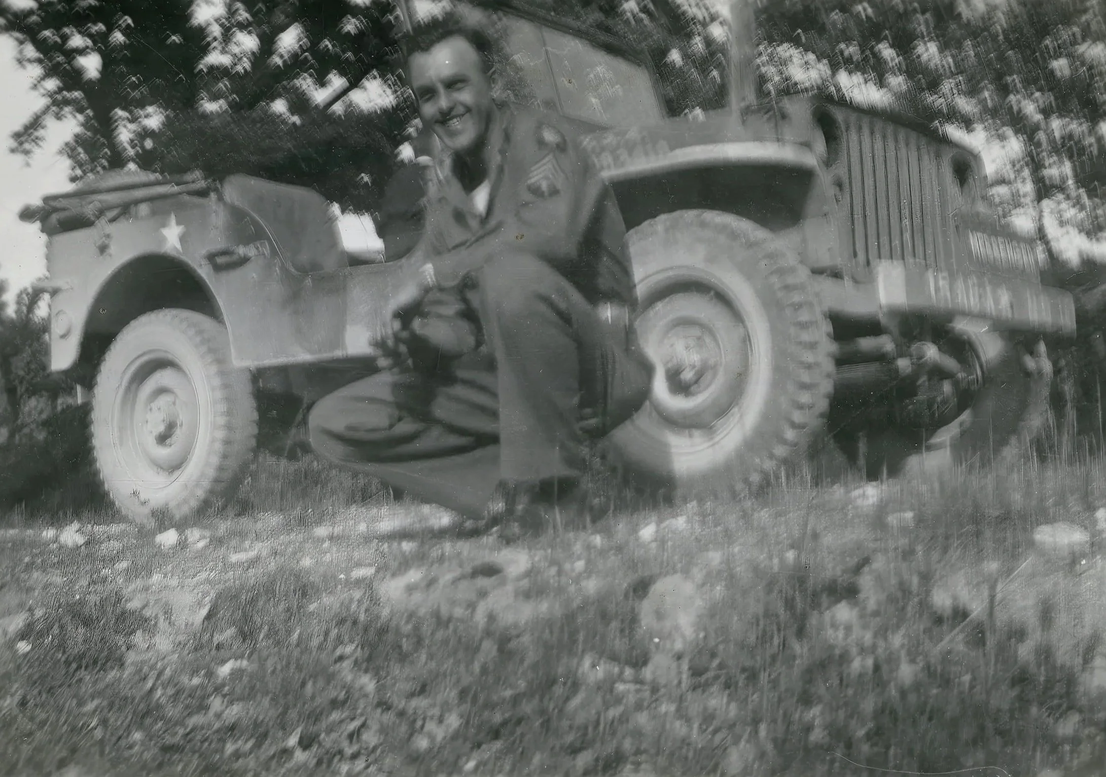 Black and white photograph of a smiling man in military uniform sitting in a vintage jeep with a star emblem on the front, in the rain, with trees reflected on the rain-splattered windshield.