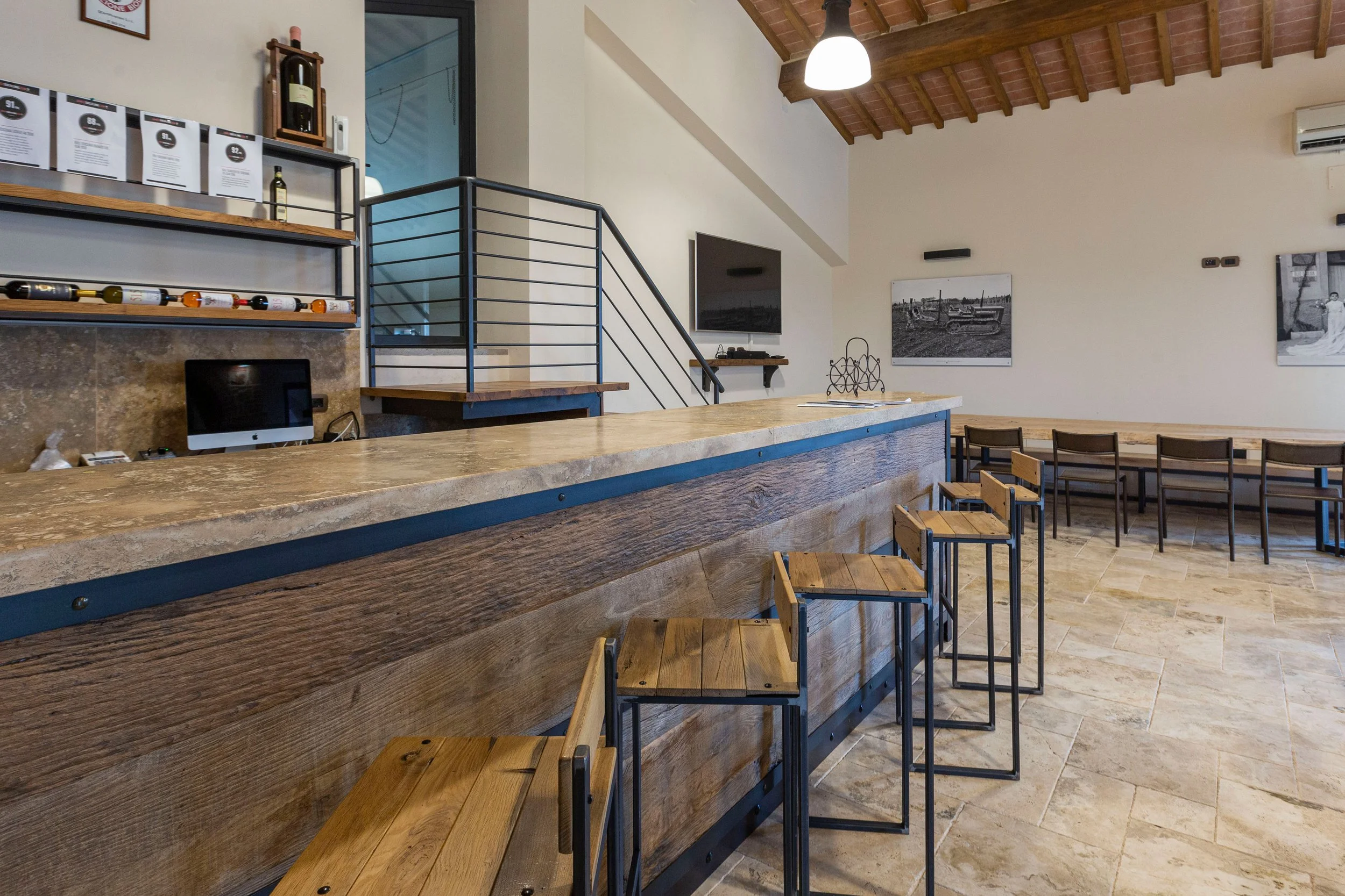 Interior of a modern restaurant or cafe with a wooden bar counter, bar stools, and a dining area with tables and chairs. The ceiling has wooden beams and a hanging light fixture. There are framed black-and-white photos on the wall, a wall-mounted television, and some decorative items and menus on the shelves.
