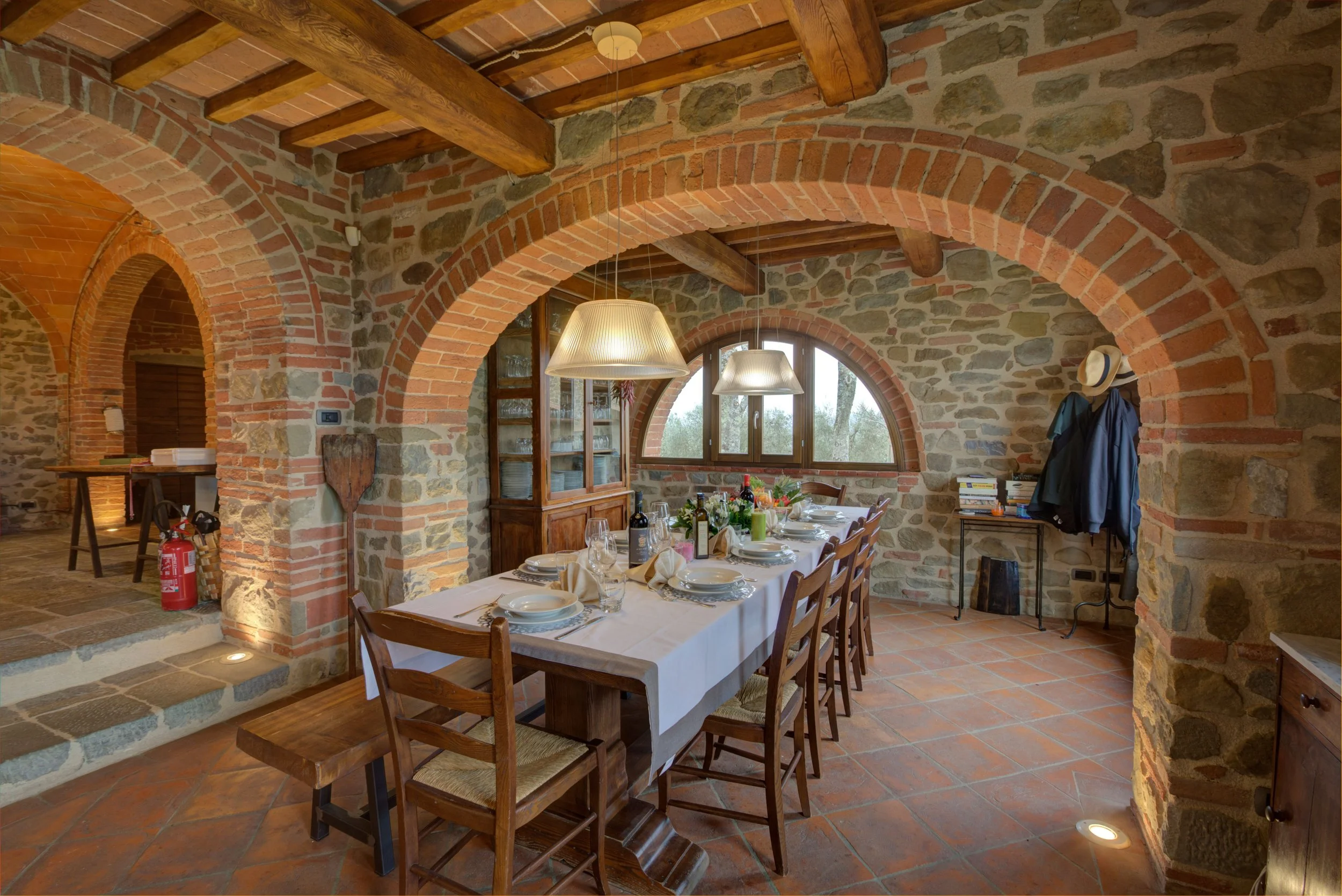 A rustic dining room with stone and brick walls and ceiling beams, set up for a meal with a long table, white tablecloth, plates, glasses, and bottles, illuminated by hanging pendant lights.