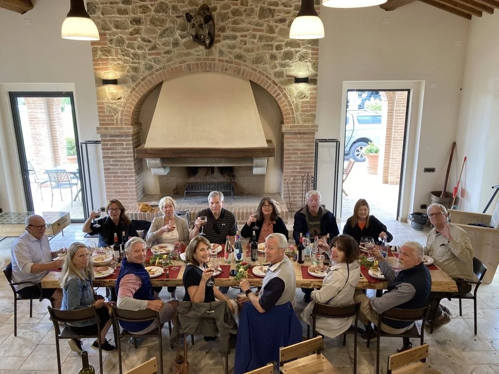 A group of fourteen people sitting around a dining table, enjoying a meal in a rustic room with a large stone fireplace, exposed wooden beams, and two open doors leading outside.