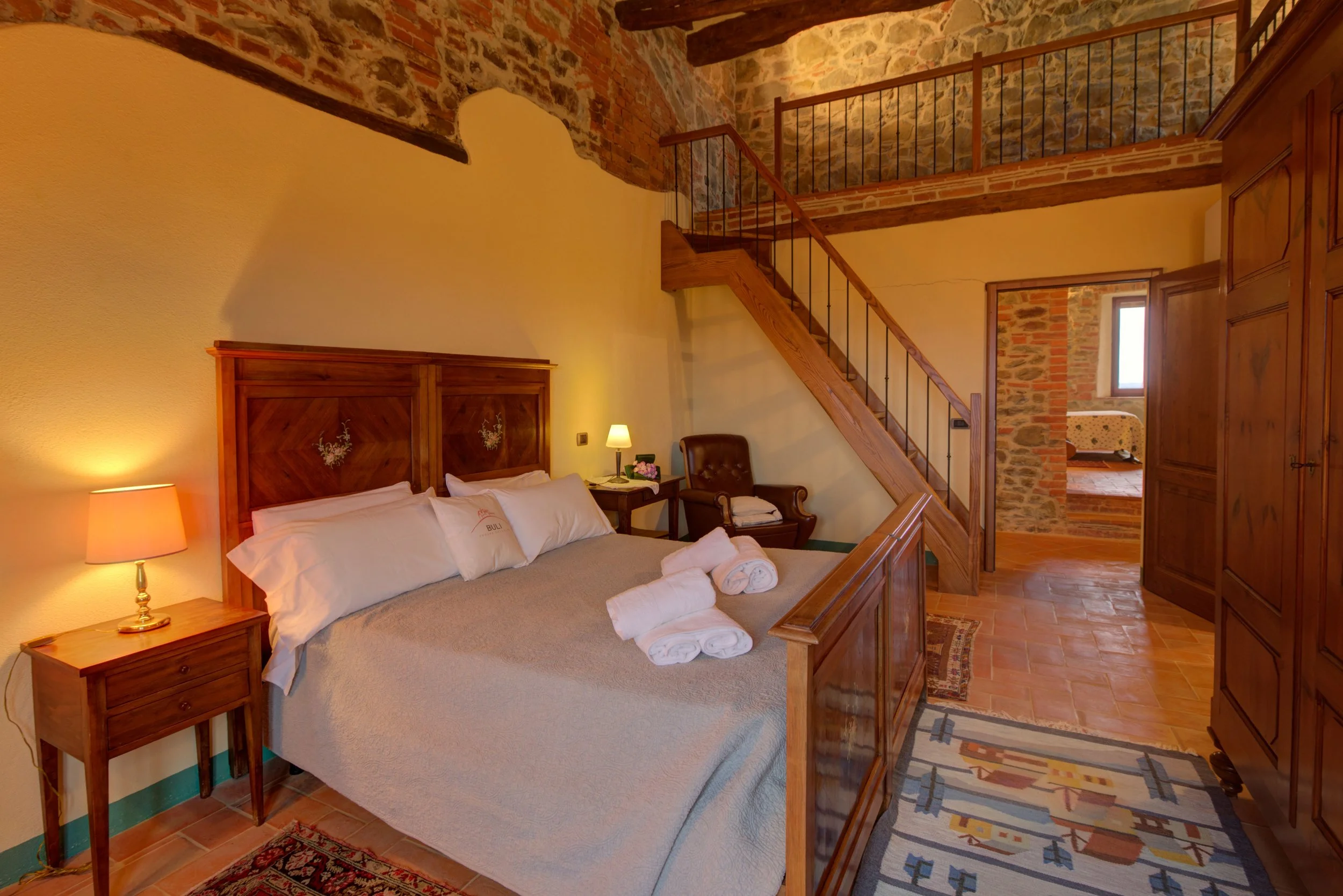 A cozy bedroom with a wooden bed, nightstand, and antique furniture, featuring exposed brick and stone walls, wooden beams, a staircase, and a window in a rustic style.