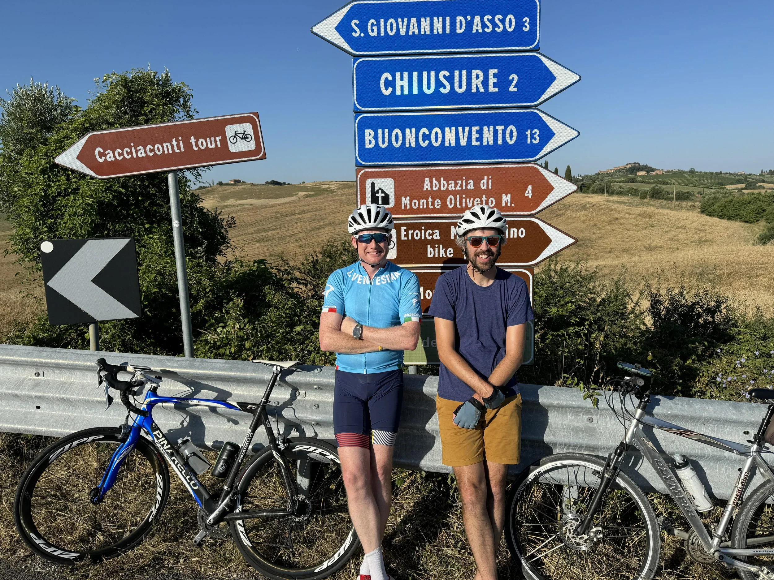 Two cyclists in front of road signs in a rural landscape, wearing helmets and cycling gear, with bicycles resting on the guardrail.
