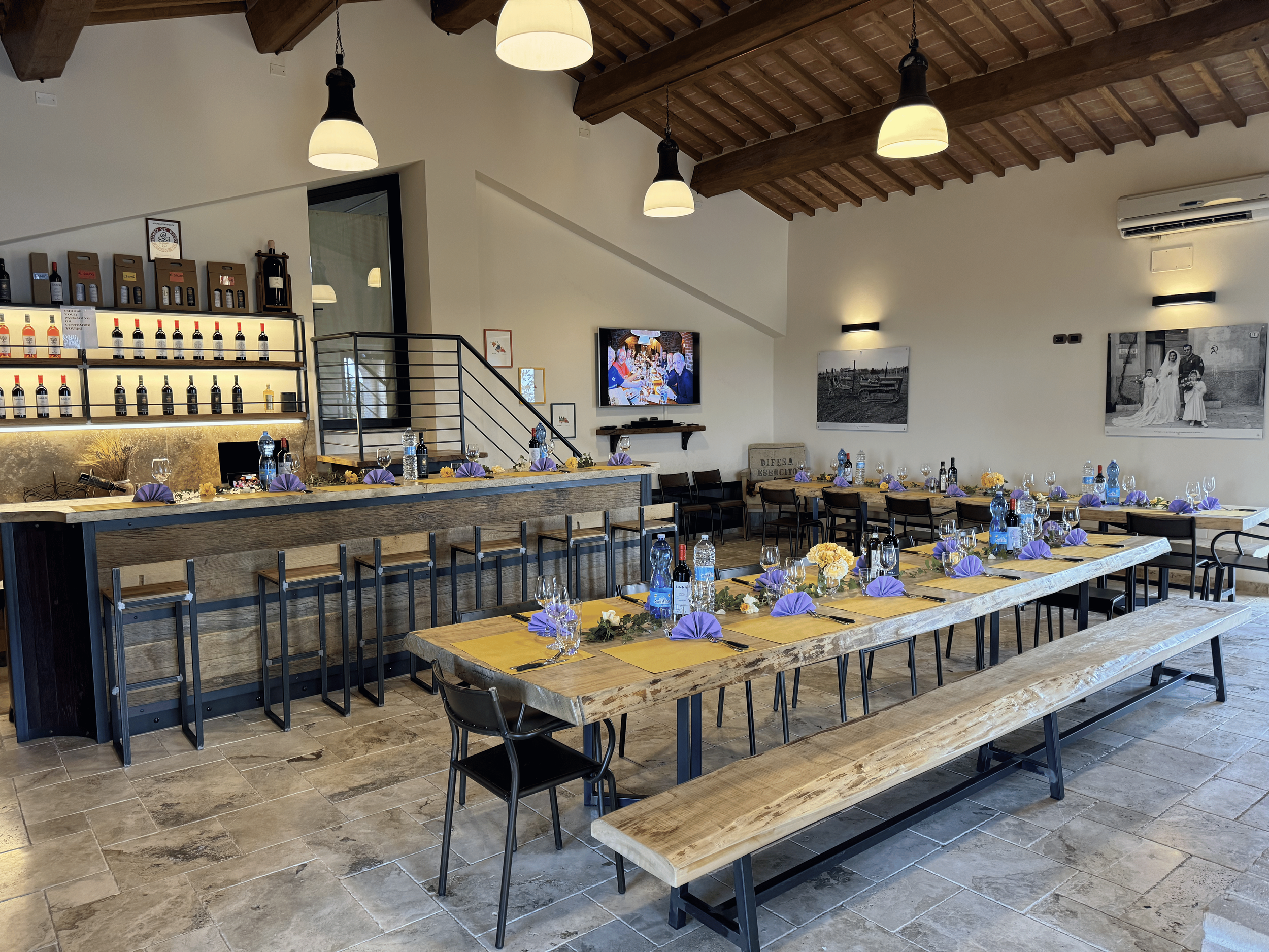 Inside a restaurant with wooden beams, hanging light fixtures, and framed black-and-white family photos on the walls. The dining area is set with tables, chairs, purple napkins, water bottles, and glasses.