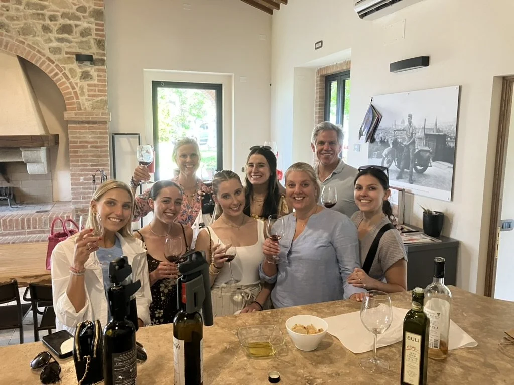 Group of eight people standing together in a restaurant, smiling and holding glasses of red wine.