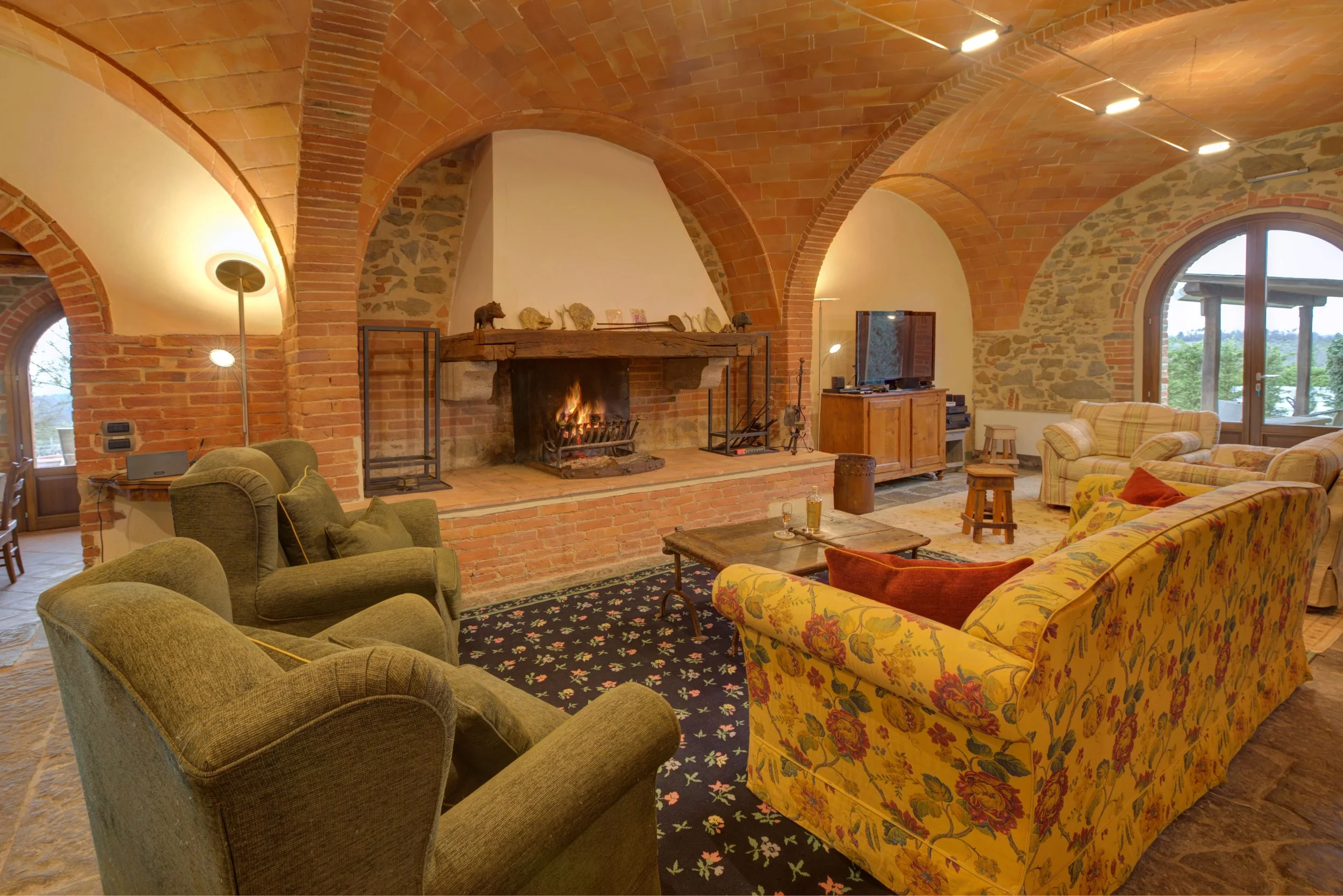 Cozy living room with brick vaulted ceilings, a fireplace, floral patterned sofa, green armchairs, wooden furniture, and large windows showing a view of trees outside.