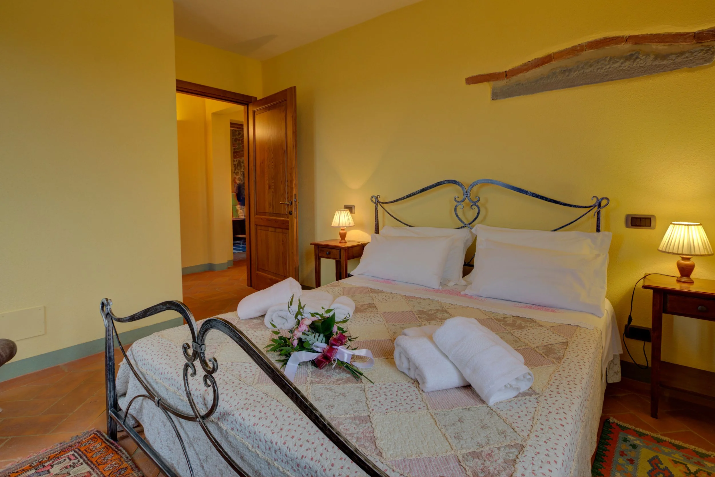 A hotel room with a bed, two bedside lamps, a wooden door slightly open showing another room, and a flower arrangement on the bed.