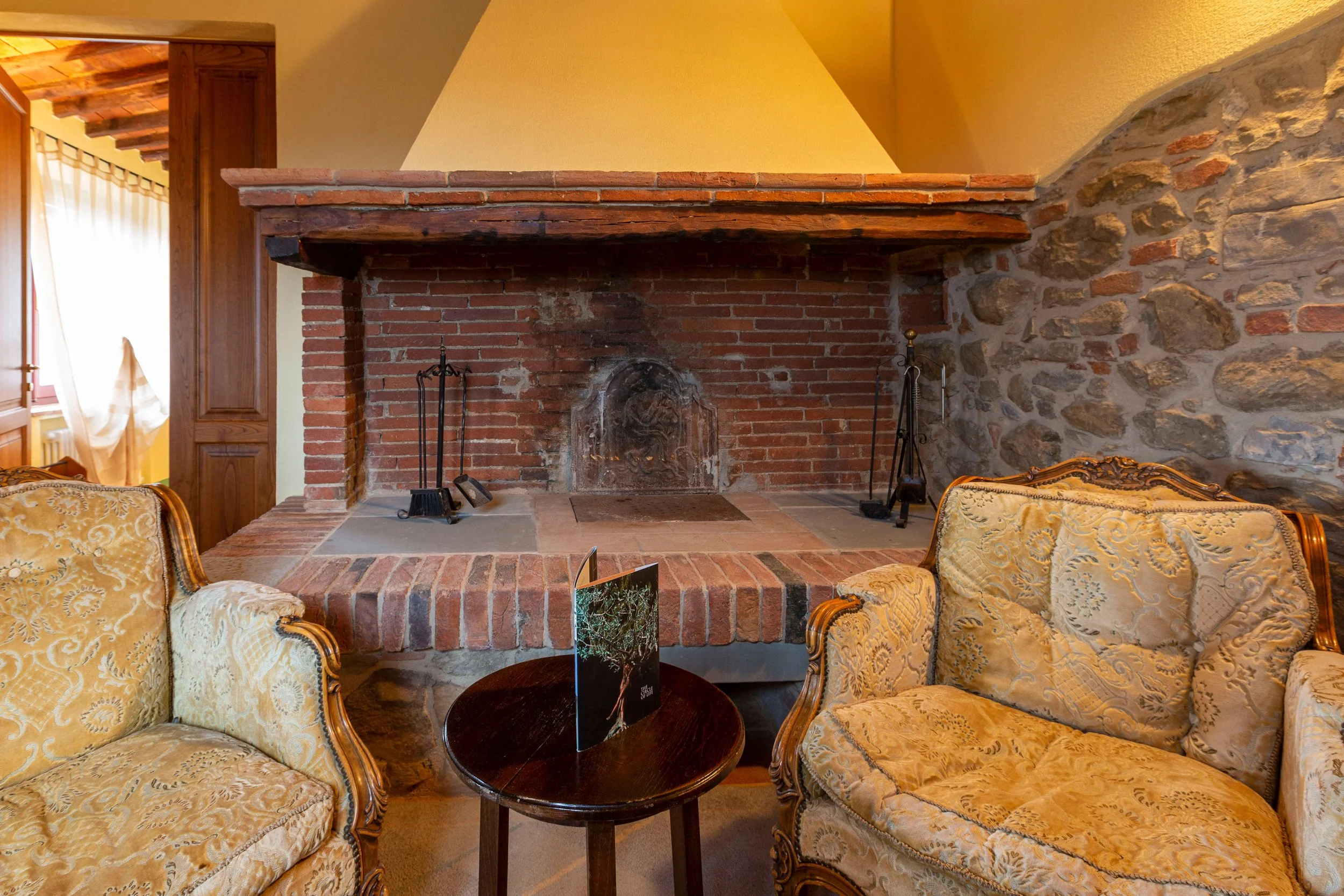 Living room with a large brick fireplace, yellow walls, and ornate yellow upholstered chairs around a dark wood table.