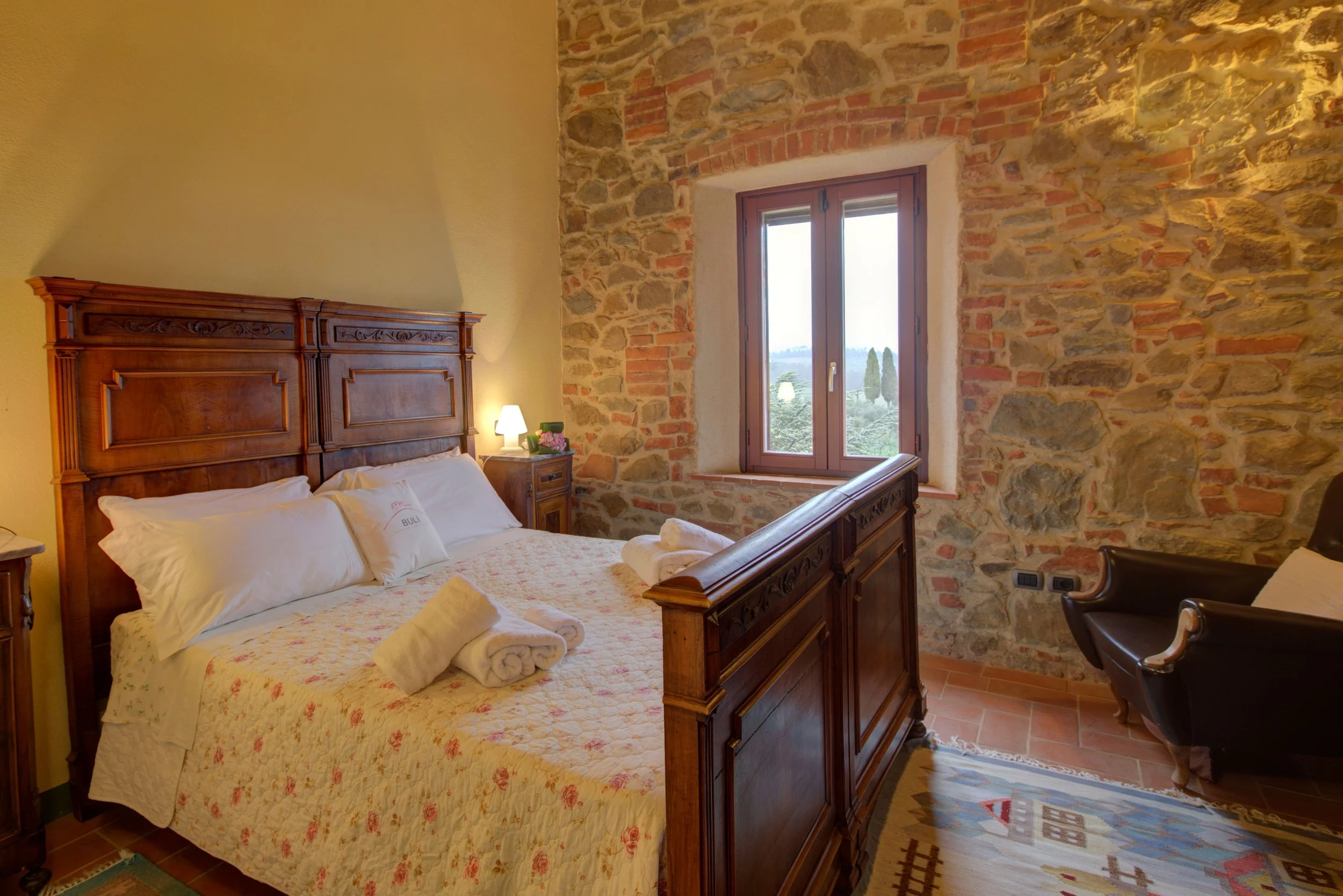 A cozy bedroom with a wooden bed frame, white bedding, and two rolled towels on the bed. There is a small side table with a lamp, a framed picture, and a window with a view of trees outside. The wall behind the bed is exposed brick and stone.