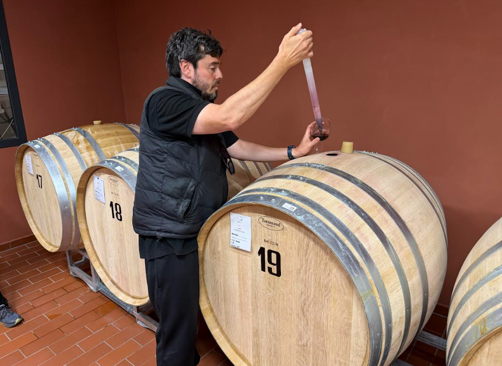Man tasting wine from a large wooden barrel in a cellar.
