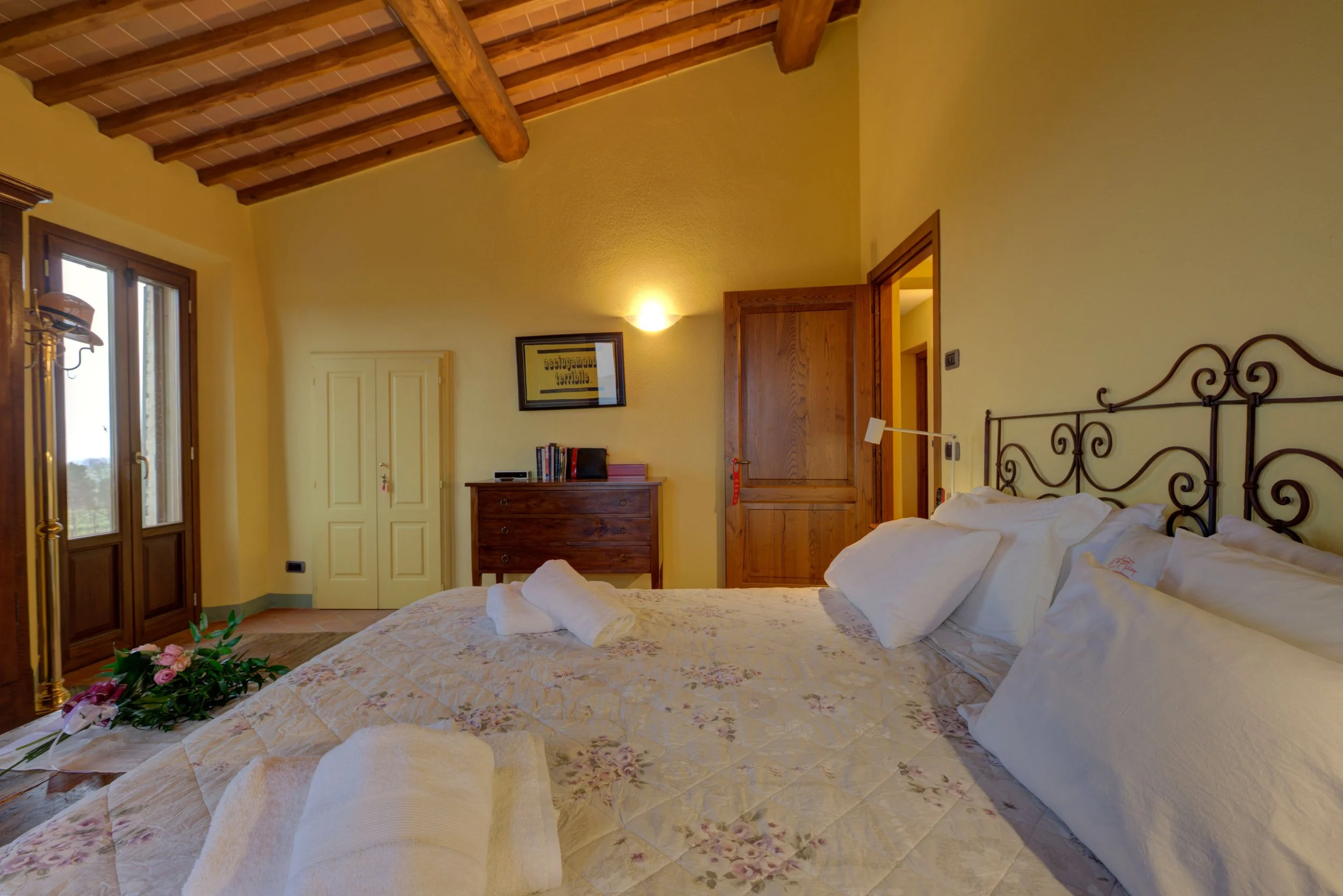 A cozy bedroom with a large bed covered in a floral quilt, with folded towels on top, and a bunch of flowers to the side. The room features wooden ceiling beams, a window letting in natural light, a dresser with books and a framed picture, and a part