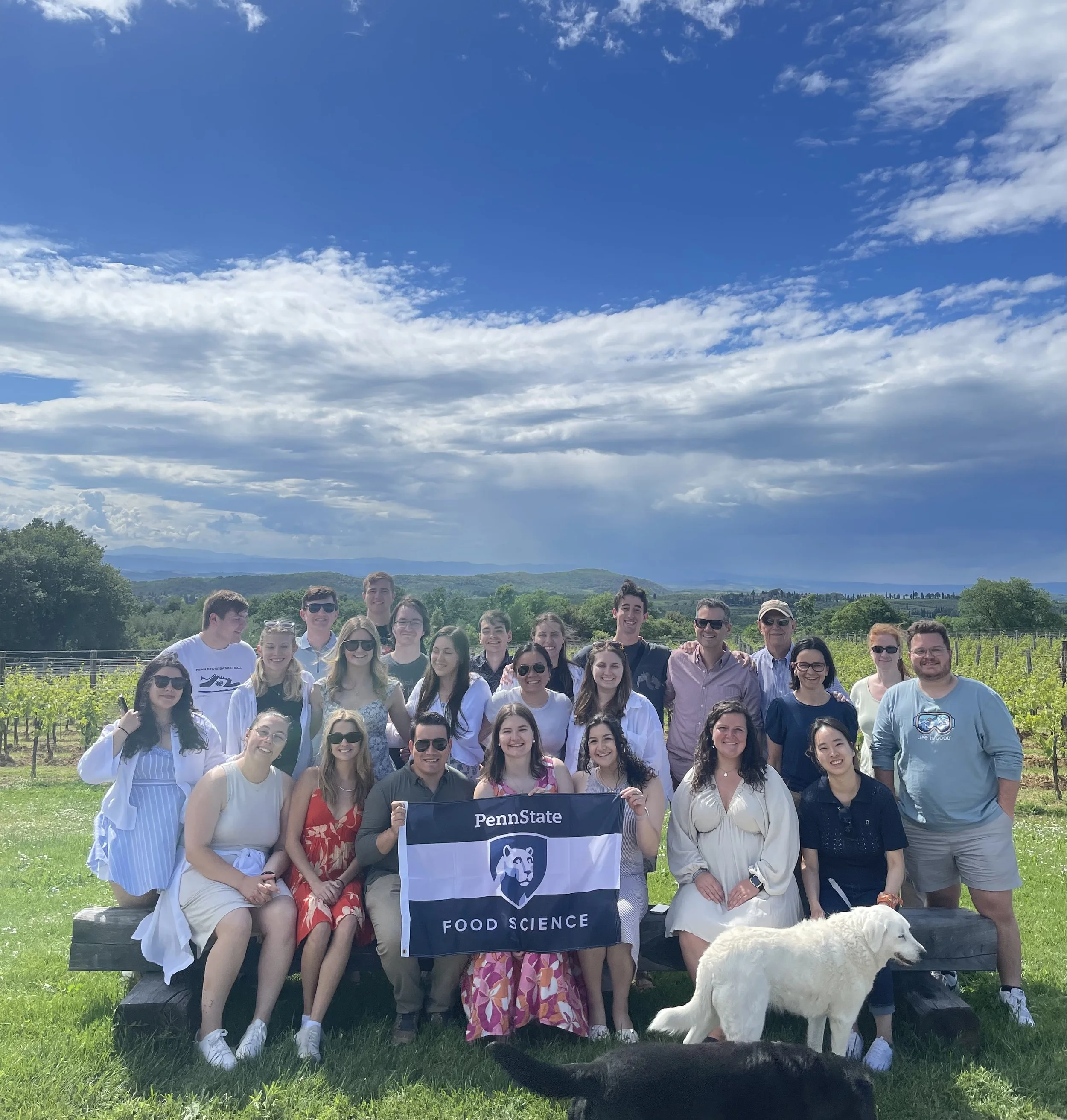 A group of people posing outdoors in front of a scenic landscape with mountains and a cloudy sky. They are holding a Penn State Food Science banner and appear to be attending a group event or celebration.