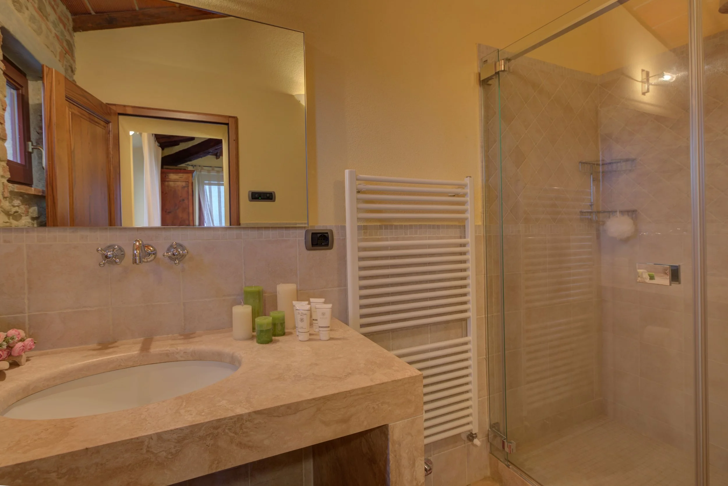 Bathroom with beige tiled countertop, mirror, white towel warmer, glass-enclosed shower, personal care products on counter, wooden door and ceiling beams.
