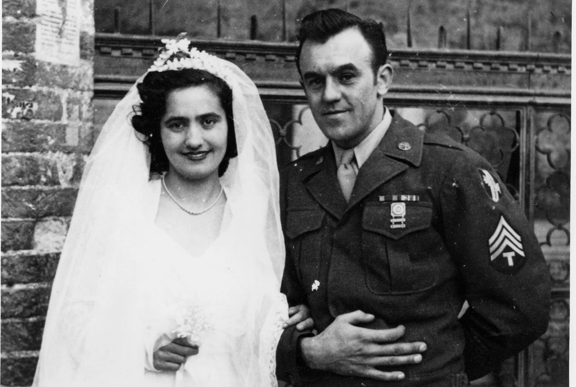 Black and white photo of a smiling bride and groom in wedding attire standing outdoors. The bride is wearing a white dress and veil, with flowers in her hair and a necklace. The groom is in a military uniform with medals and patches, standing with his hand on his chest. They are in front of a brick and wood fence.